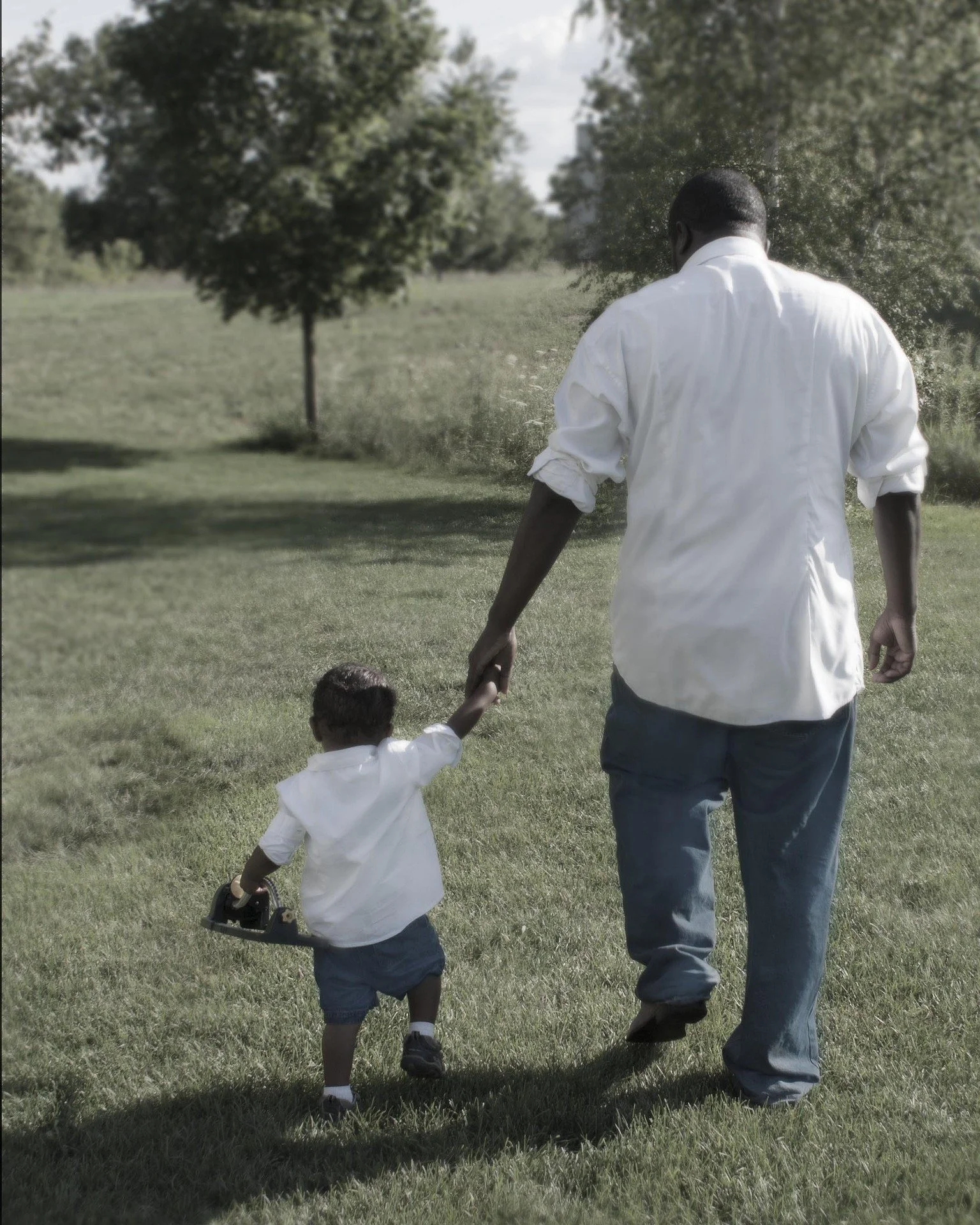 A man and a boy walk hand in hand across a grassy park. The man wears a white shirt and blue jeans, and the boy wears a white shirt and dark shorts. The girl holds a toy truck.