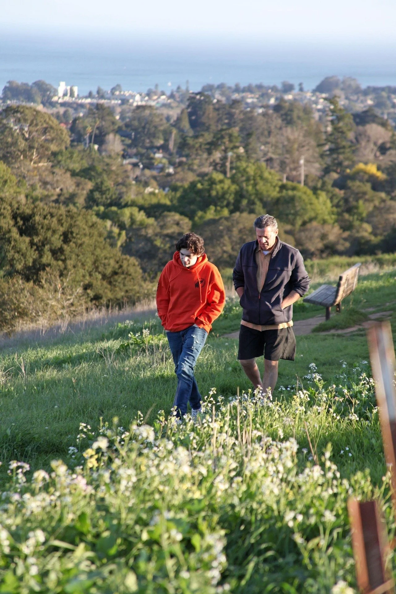 Two men walking in a grassy field with trees and a town in the background, one wearing a red hoodie and the other wearing a black jacket.