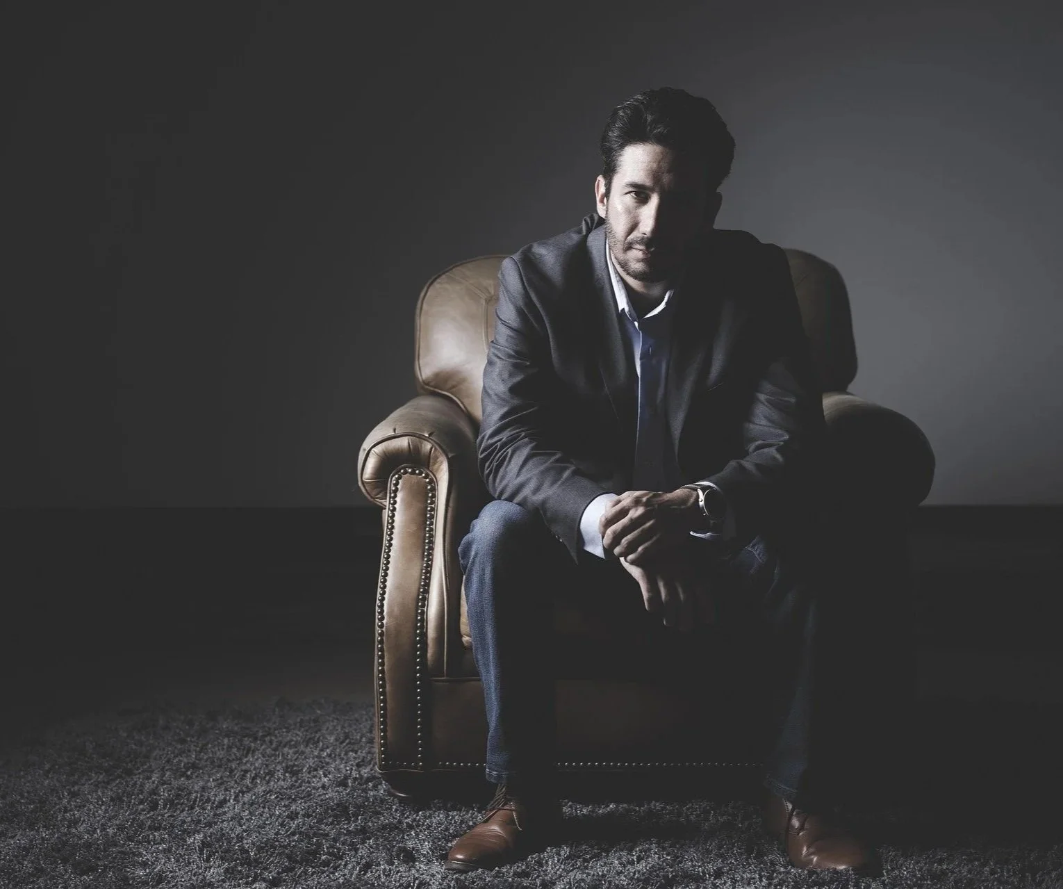 A man sitting on a brown leather armchair in a dimly lit room with a dark gray background, dressed in a suit and looking serious.