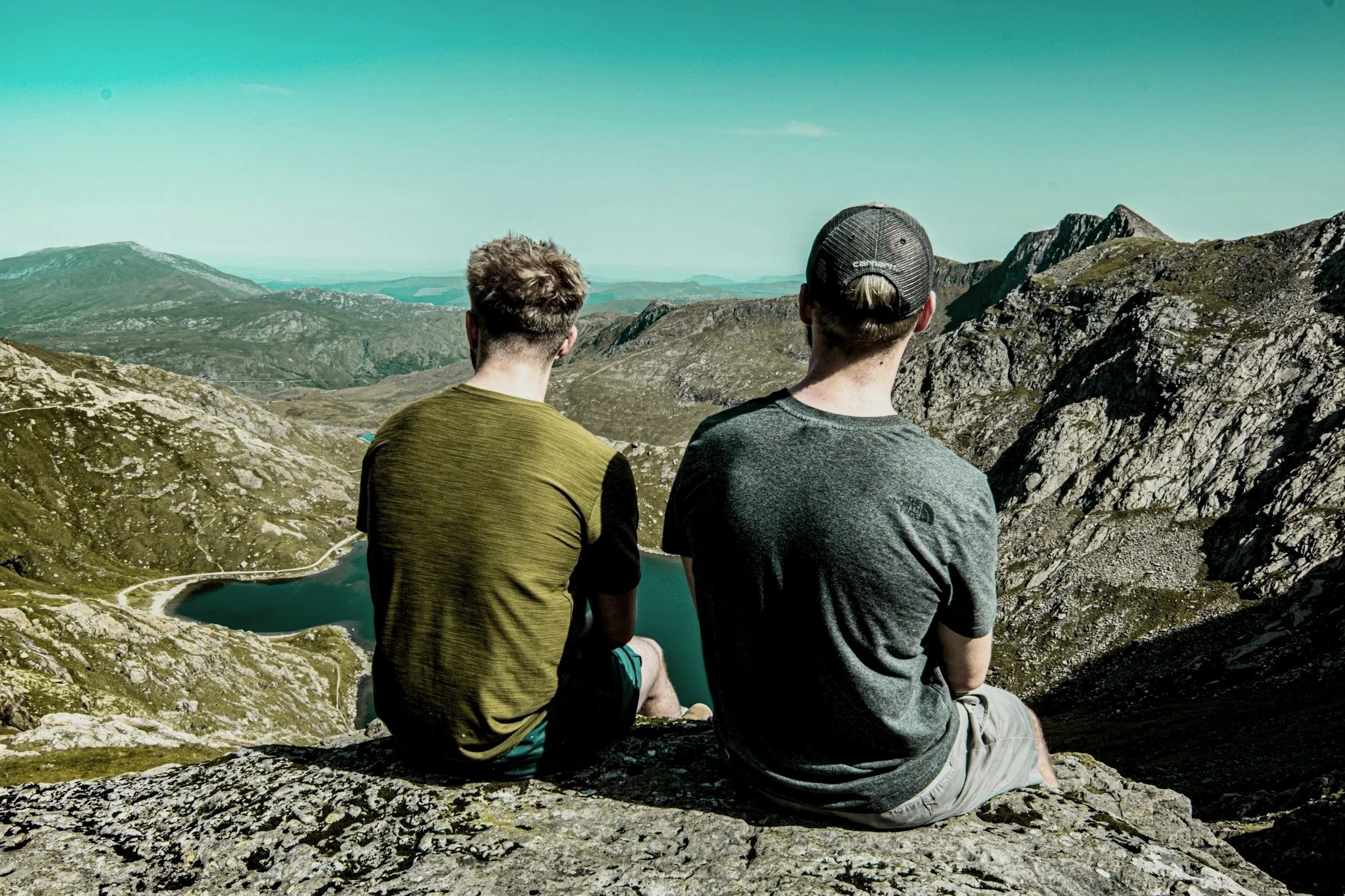 Two men sit on a rocky ledge overlooking a mountain landscape with lakes and rolling hills, under a clear sky.