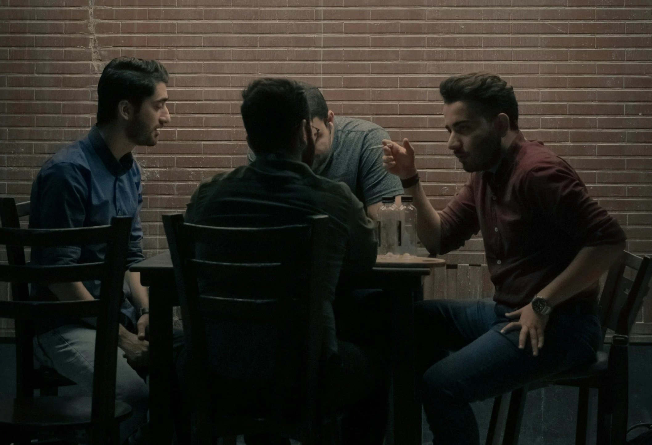 Four young men sitting at a table, engaged in conversation, with a brick wall in the background.