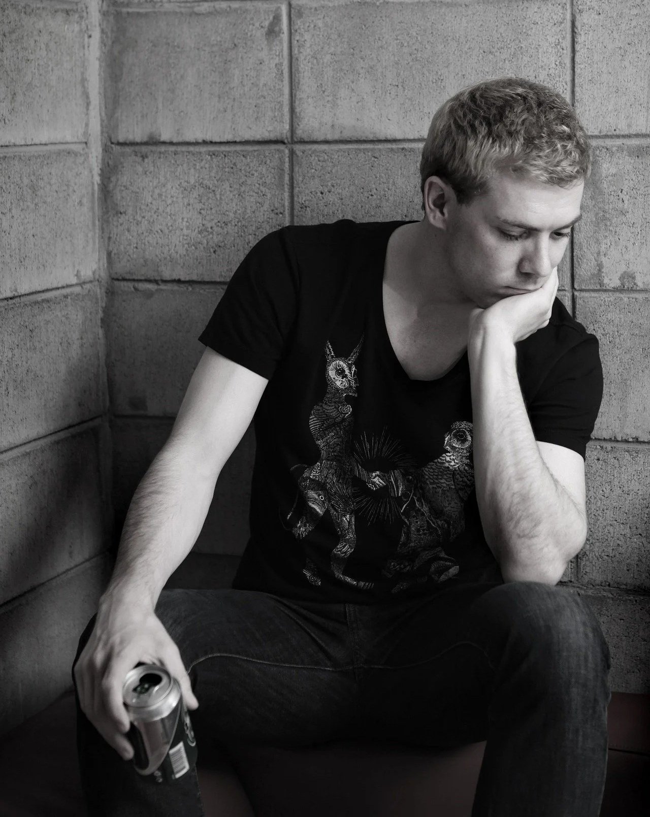 A young man with curly hair sitting against a brick wall, holding a soda can in one hand, resting his chin on his other hand, looking down with a thoughtful expression.