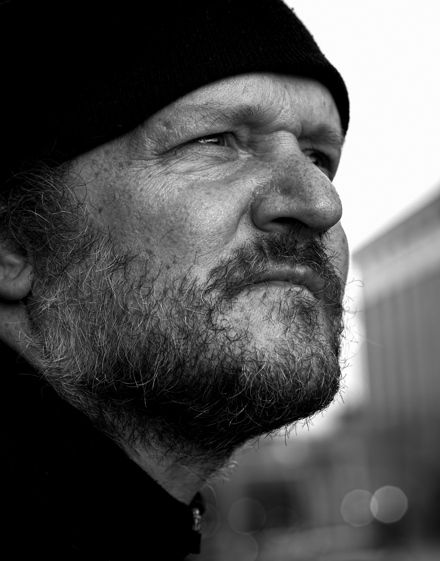 Close-up of a man's face in profile, black and white, wearing a black beanie, with a beard and mustache, looking to the right.