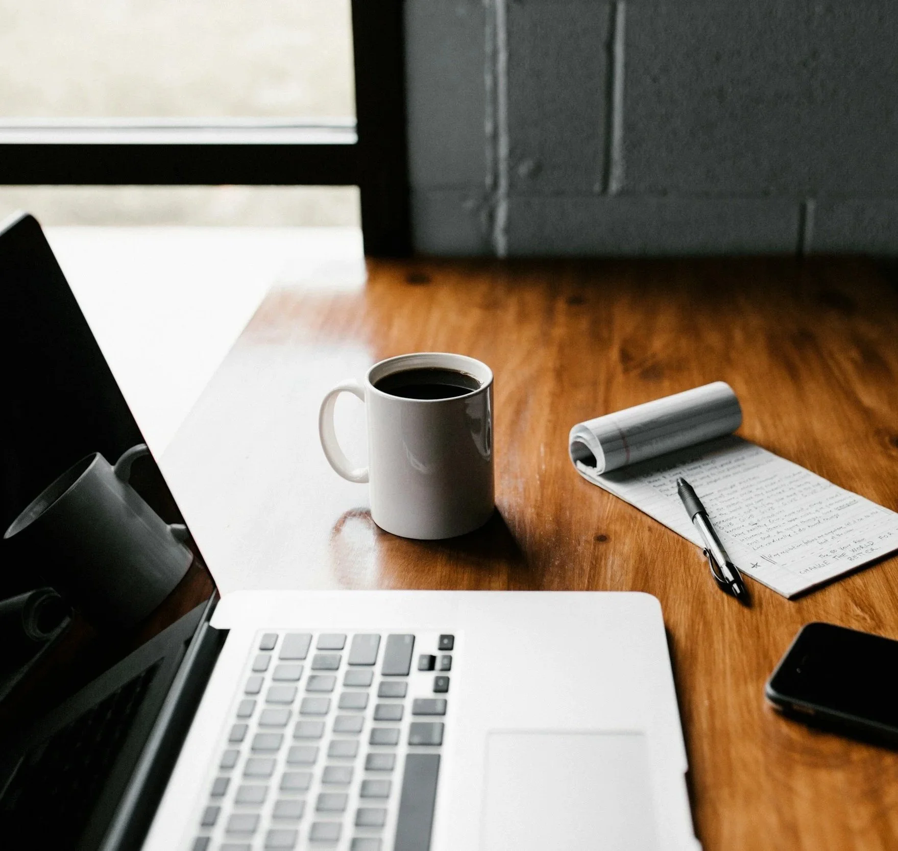 Start of a workday desk with a coffee mug, a rolled-up newspaper, a notebook, a pen, a laptop, and a smartphone on a wooden table, with a window in the background.
