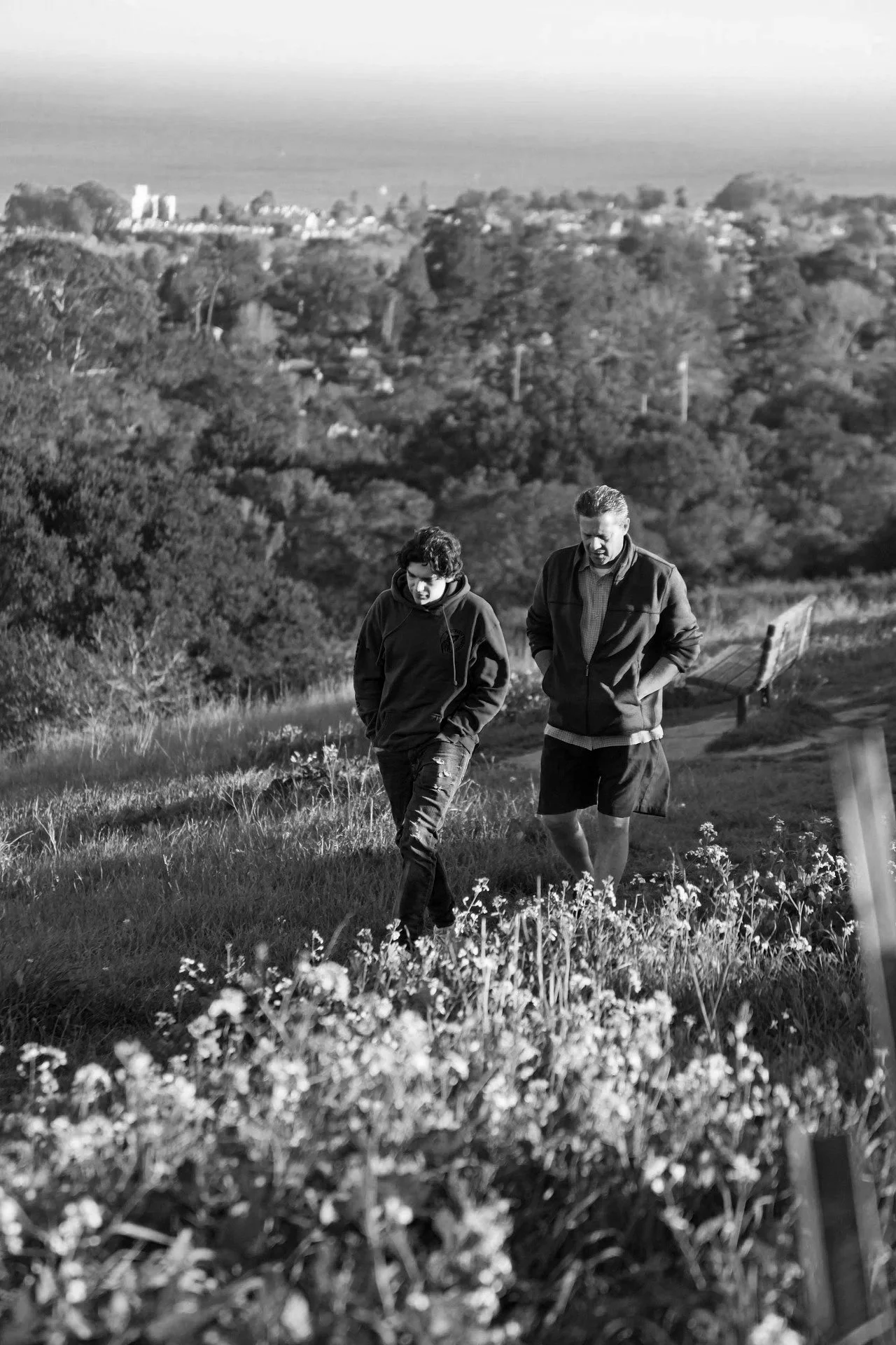 Two people walking through a grassy area with flowers in foreground, trees and a cityscape in background, black and white photo.