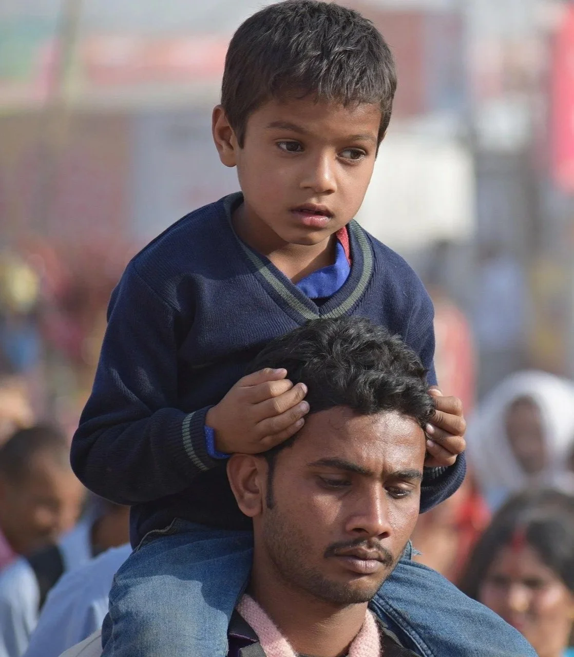 A boy sitting on a man's shoulders, holding his head and looking contemplative, with a blurred crowd in the background.