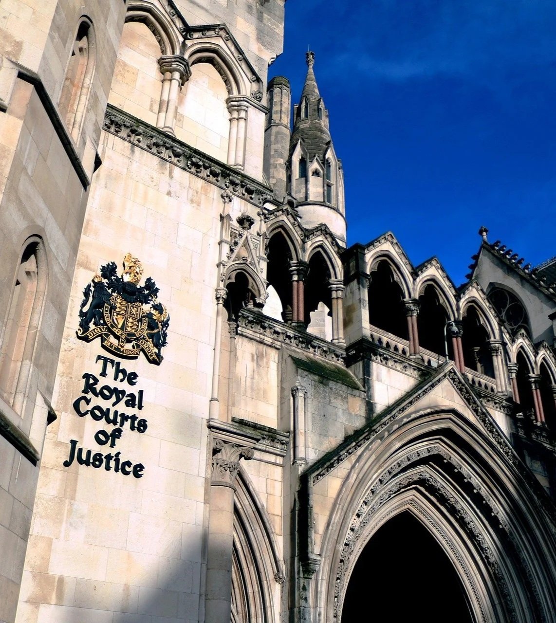 The entrance to the Royal Courts of Justice with Gothic architectural style featuring pointed arches and ornate detailing. A shield and crest emblem is visible on the building wall, with clear blue sky in the background.