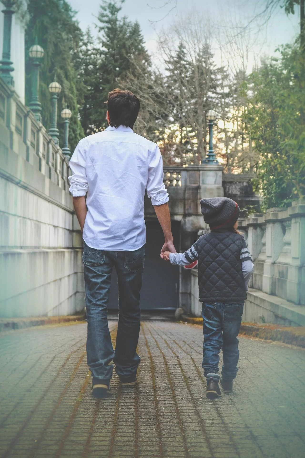 A man and a young boy walking hand in hand on a cobblestone path surrounded by trees.