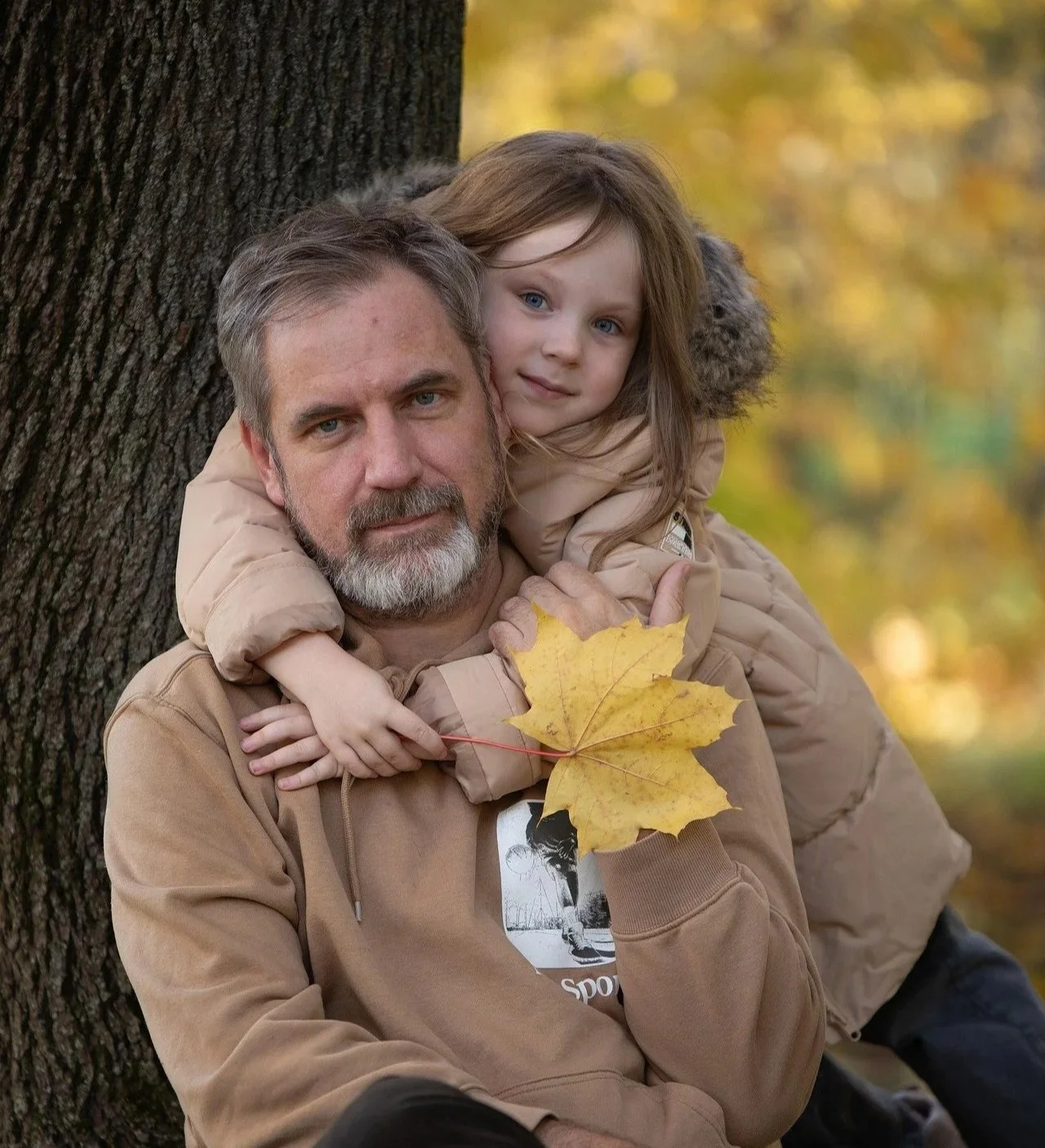 An older man with gray hair and a beard holding a young girl with long brown hair against a tree in an autumn park, both wearing beige jackets, with fallen yellow leaves.