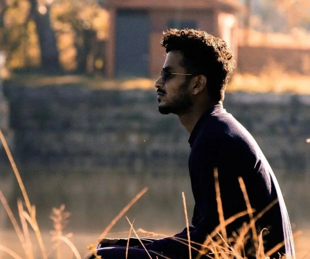 A man with curly hair and sunglasses sitting outdoors near a lake, with dry grass in the foreground and a structure in the background, during what appears to be sunset.