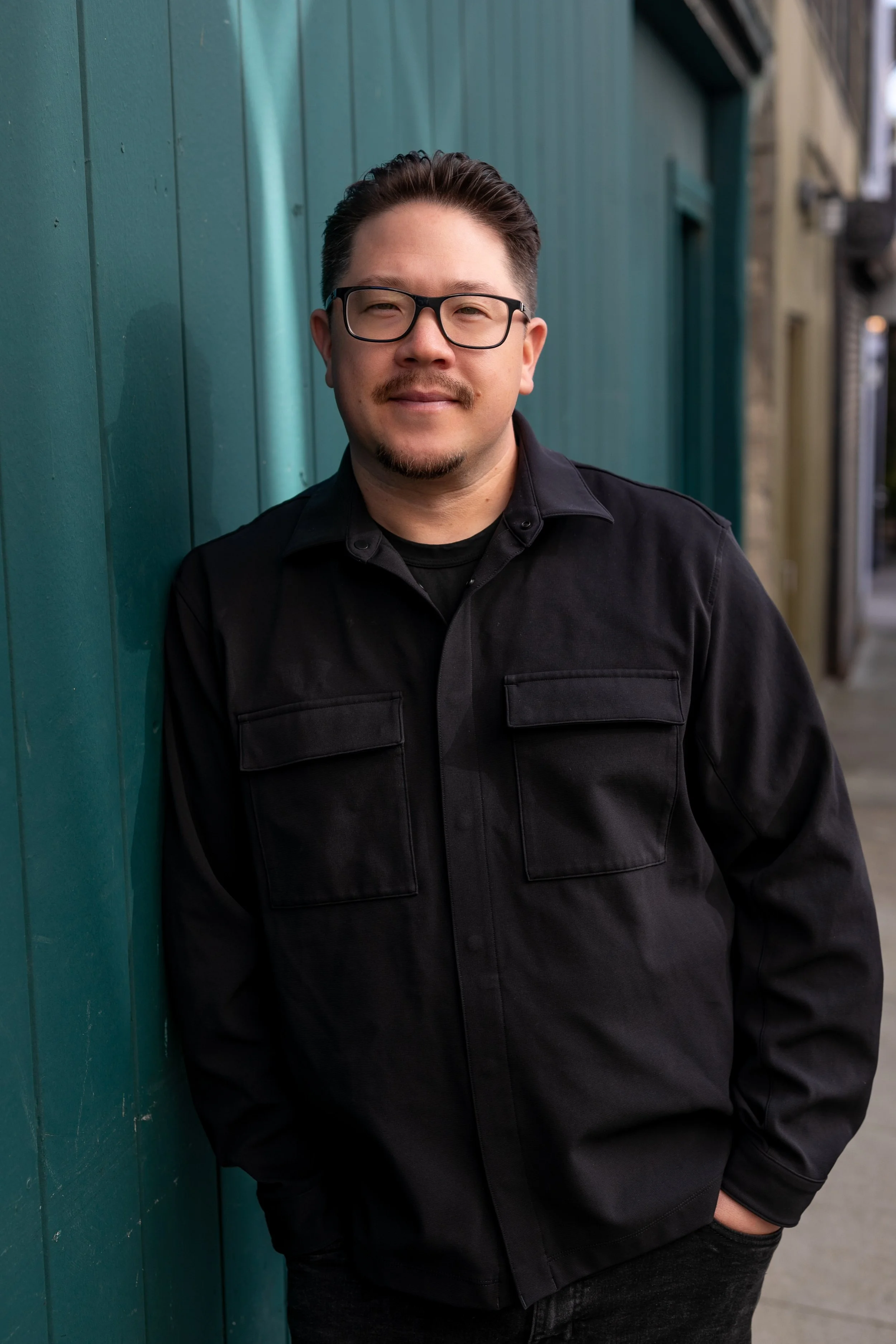 Christian wearing glasses and a black jacket standing against a teal-colored wooden wall on an urban street.
