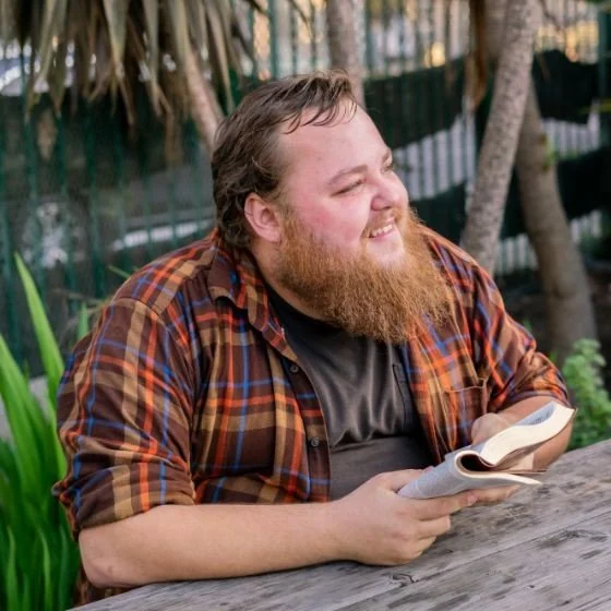A bearded man in a plaid shirt sitting outdoors reading a book and smiling.