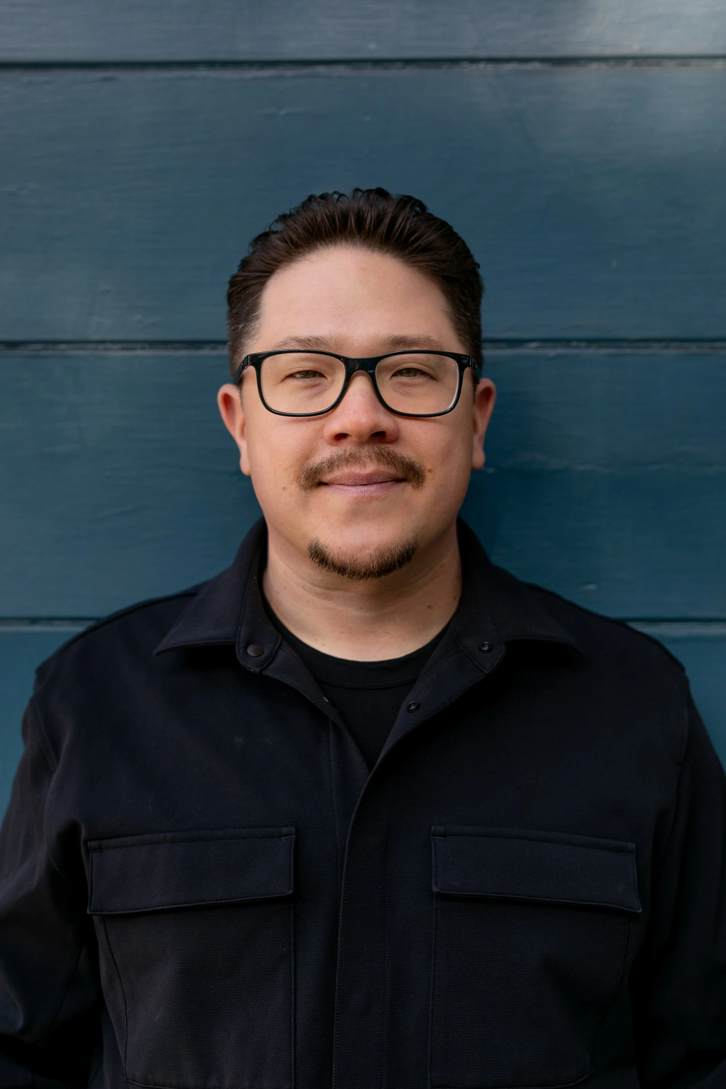 A man with glasses and facial hair standing against a blue wooden wall.