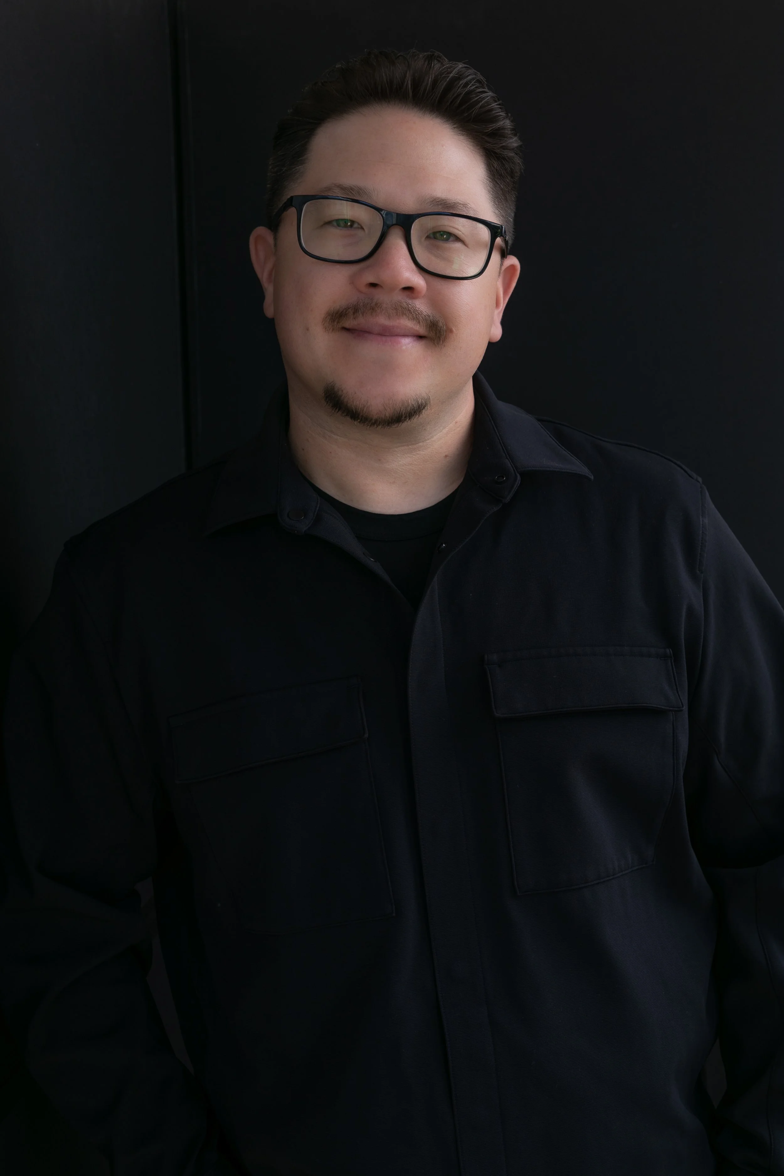 A man with glasses and a goatee in a black shirt standing against a black background.
