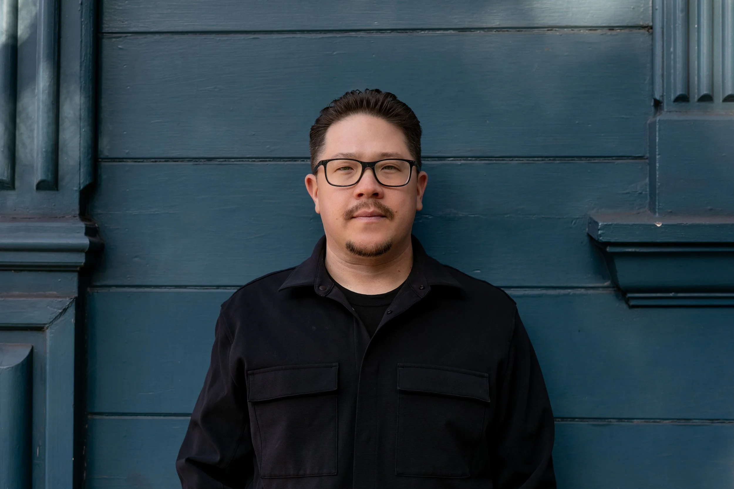 A man with glasses and a goatee standing in front of a blue wooden wall.