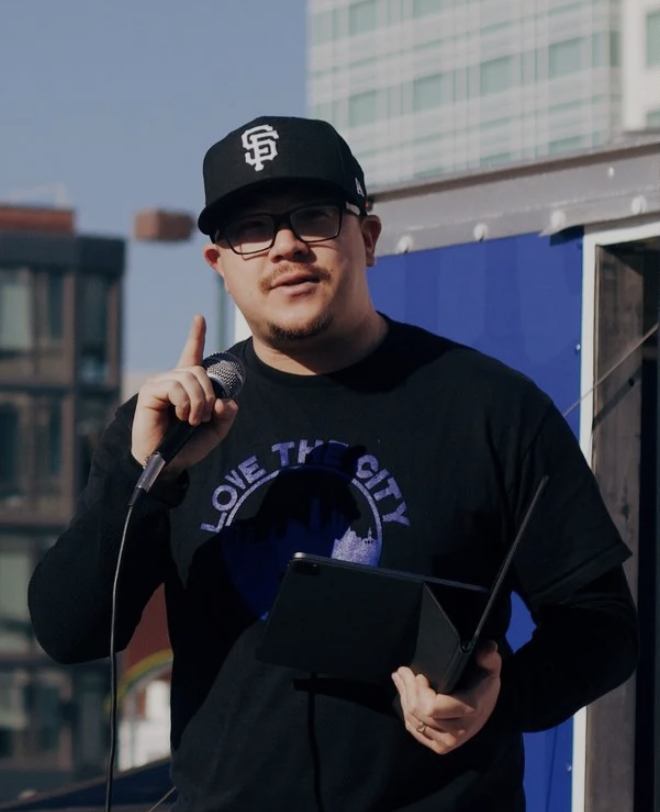 A young man wearing glasses and a San Francisco Giants cap is holding a microphone and a box, speaking at an outdoor event in a city setting.