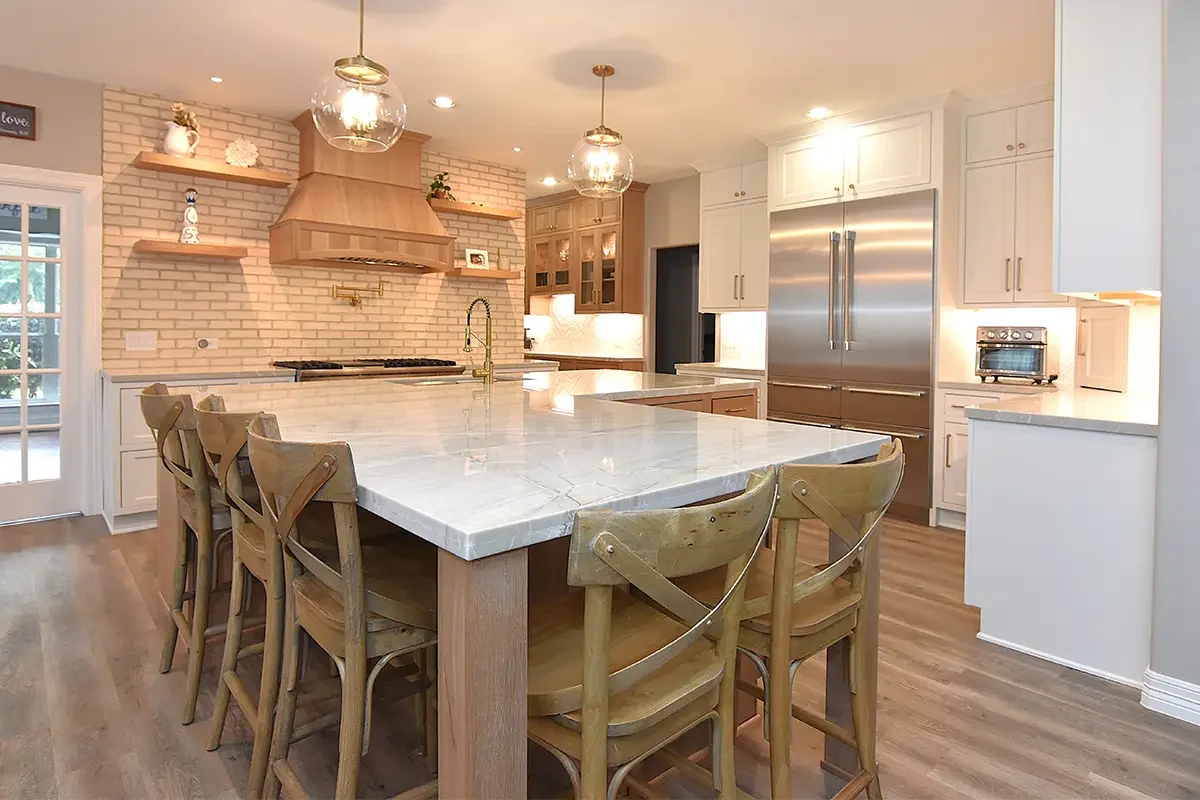 Modern kitchen with a large marble island, wooden chairs, white cabinets, stainless steel refrigerator, brick accent wall, and glass pendant lights.