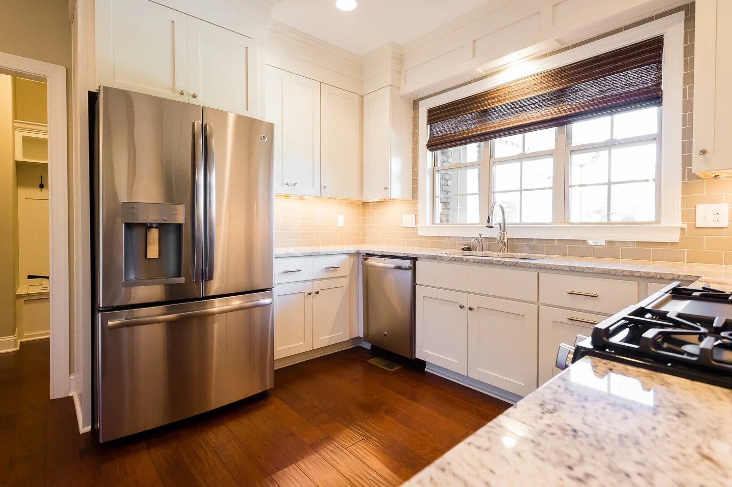 Modern kitchen with stainless steel refrigerator, dishwasher, gas stove, white cabinets, beige tile backsplash, large window with brown blinds, granite countertop, and hardwood floors.