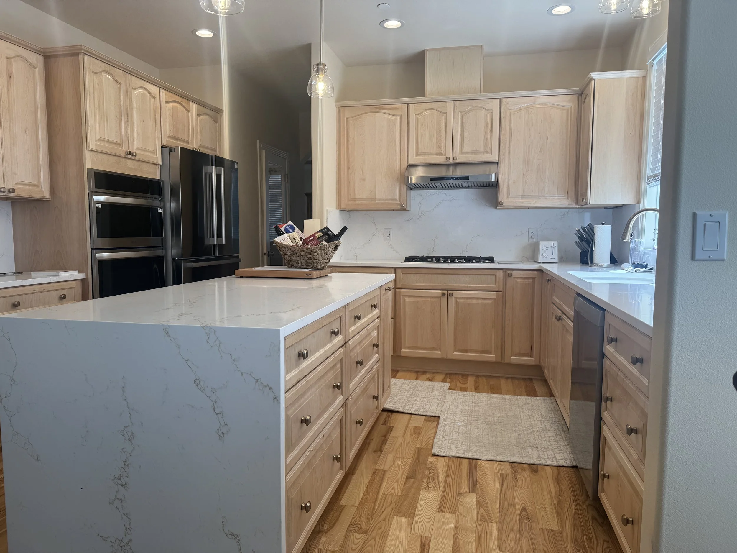 A modern kitchen with light wood cabinets, white marble countertops, stainless steel appliances including a double oven and refrigerator, a kitchen island with a basket of wine bottles, hardwood floor, and a window near the sink.
