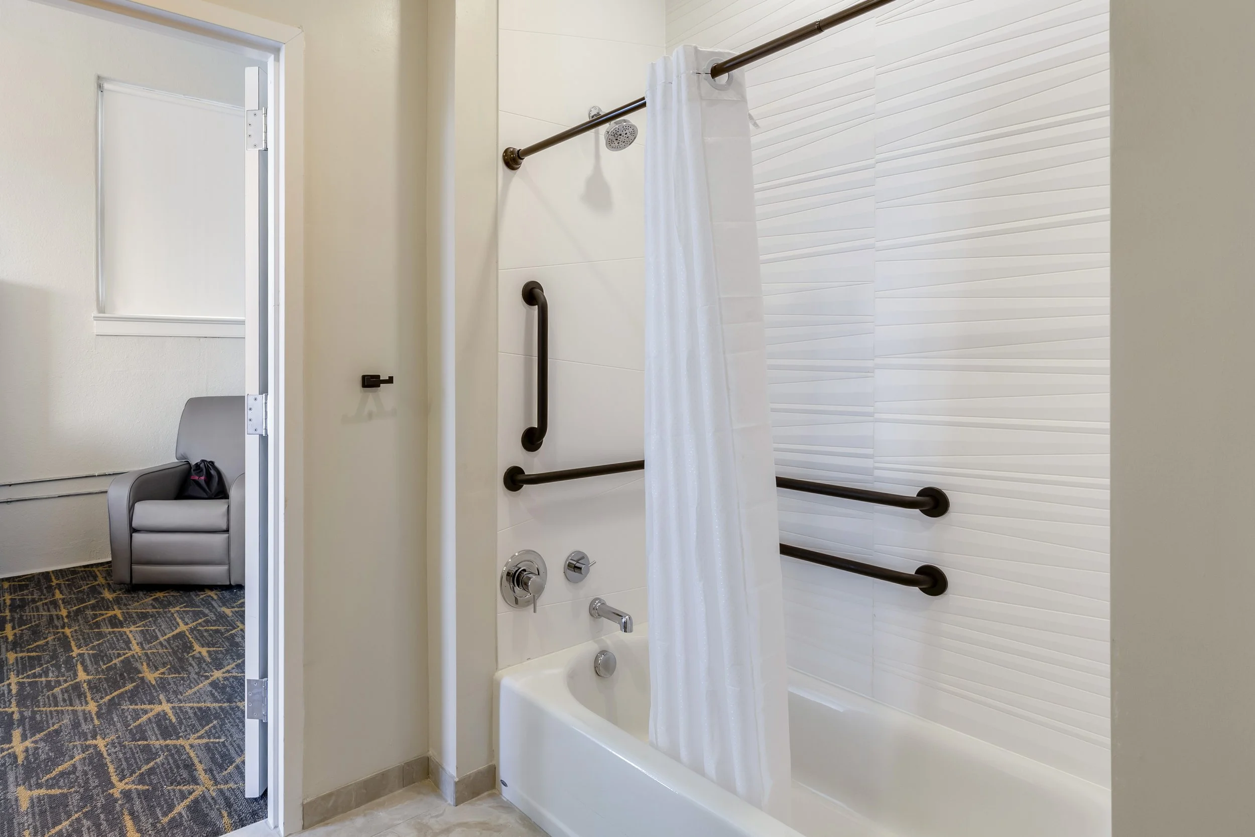 Accessible bathtub with grab bars and a shower curtain in a bathroom, with an adjacent room featuring a gray armchair and patterned carpet.