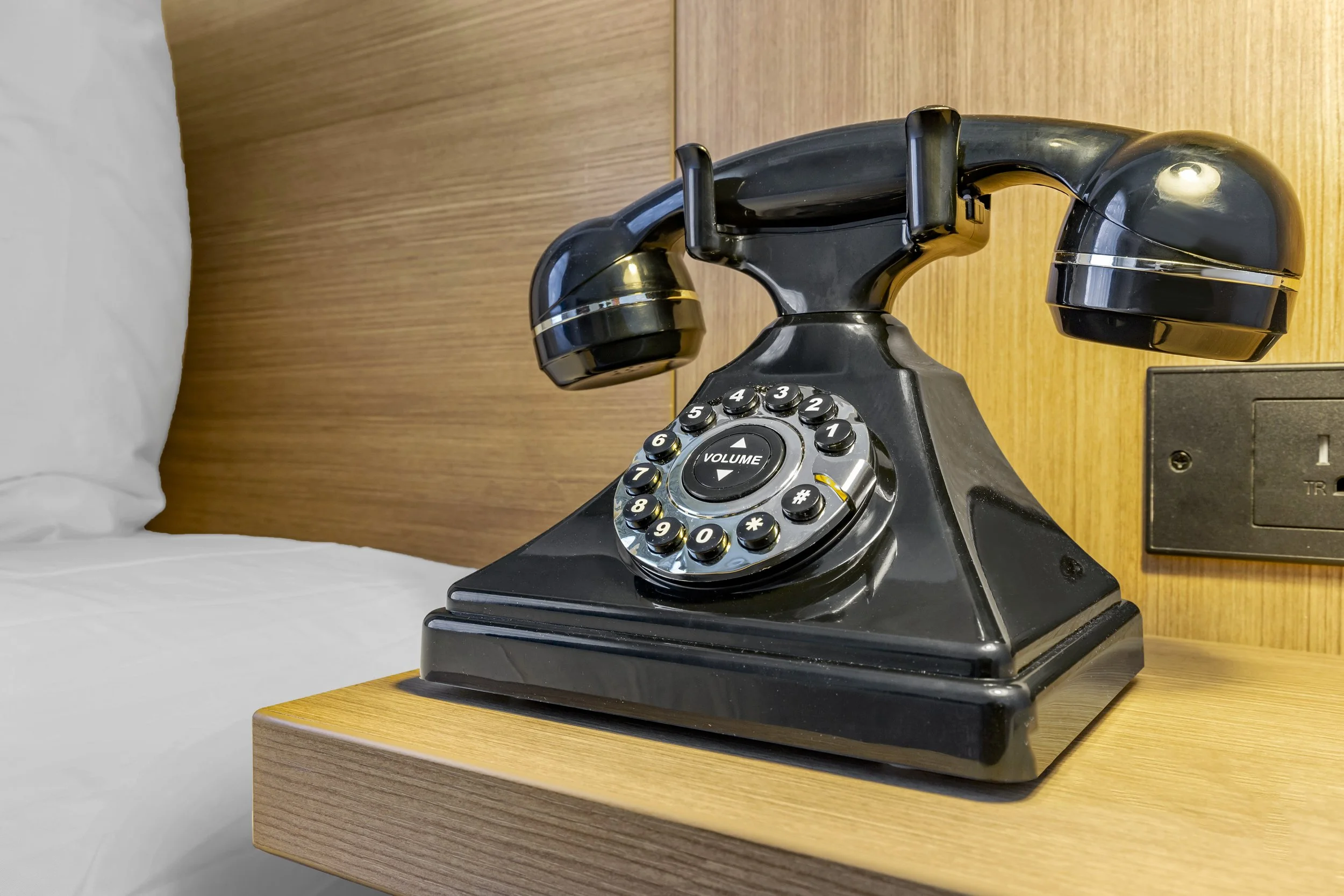 A vintage black rotary phone on a wooden bedside table in a hotel room.