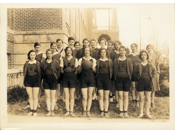 Black-and-white photo of a girls' basketball team sitting and standing outdoors in front of a building, some wearing uniforms and sneakers, with a basketball in the center.
