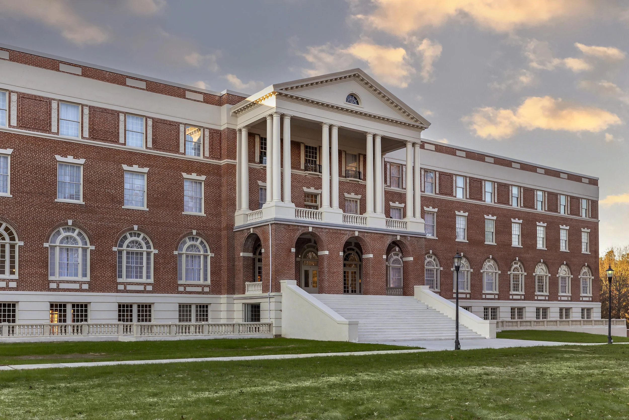 A large brick building with classical architecture features, including columns and arched windows, set against a partly cloudy sky during sunset.