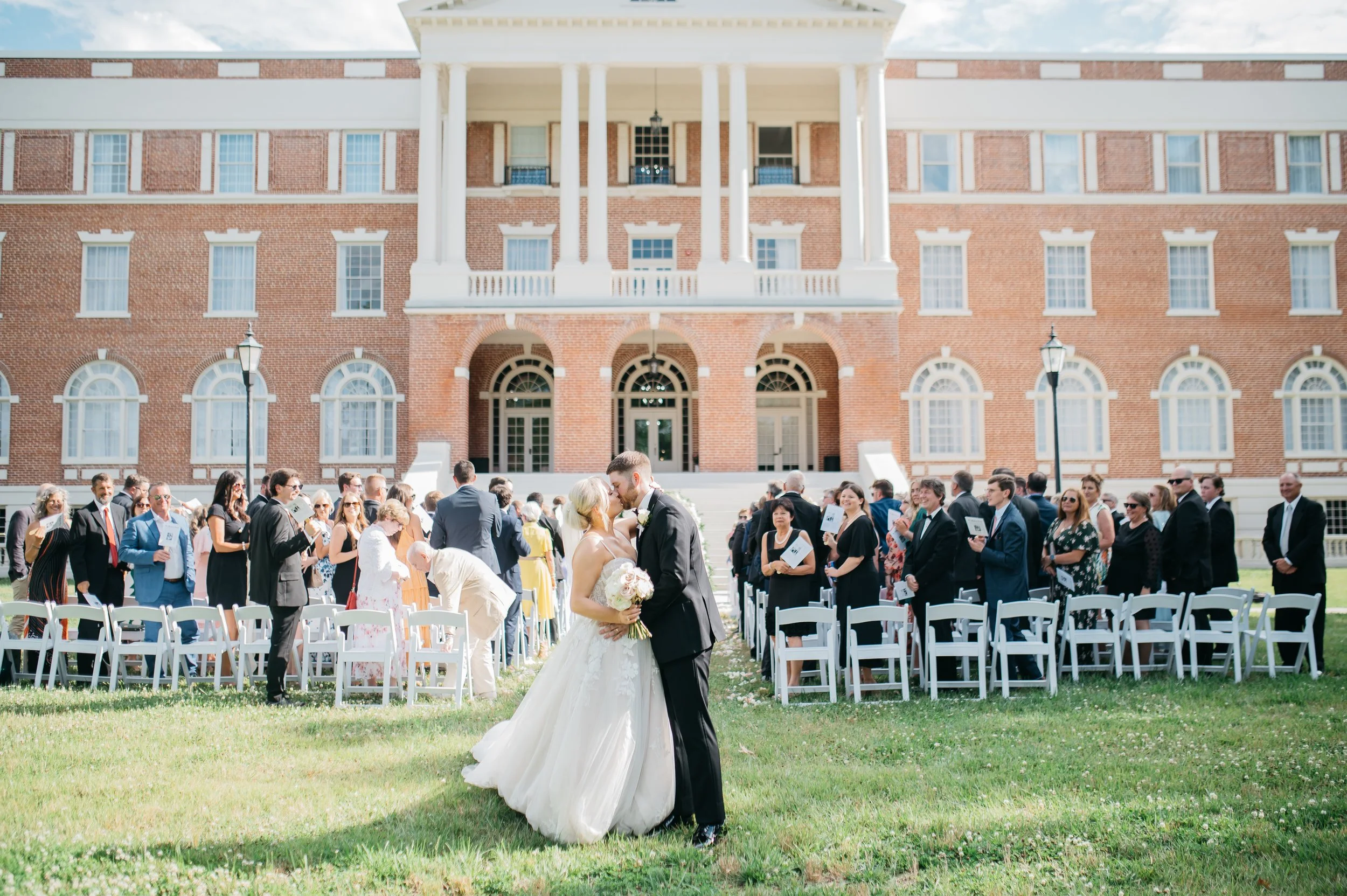 A bride and groom share a kiss in front of their wedding guests seated in white chairs on a lush green lawn, with a large brick building with arched windows and columns in the background under a blue sky.