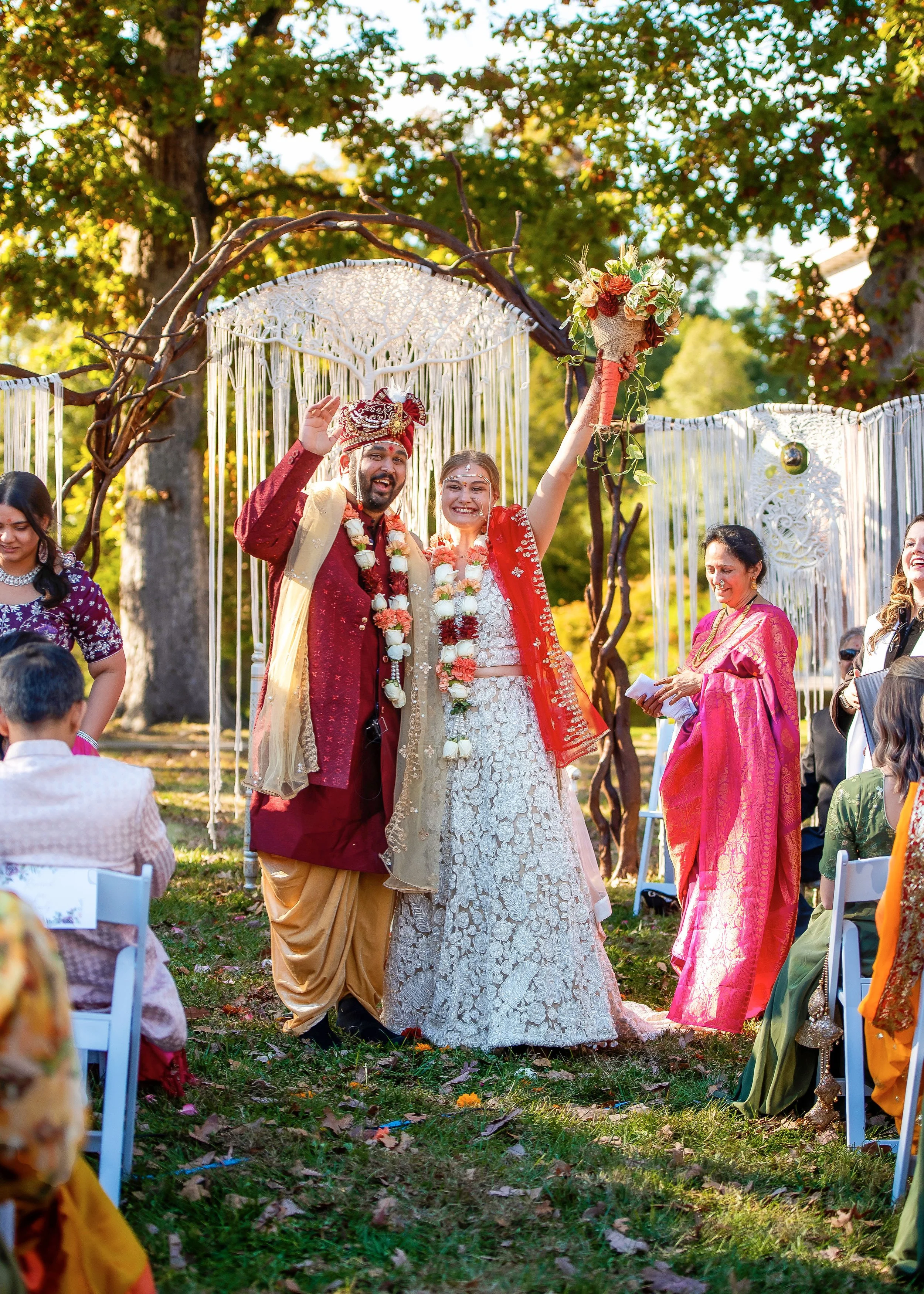 A newlywed couple in traditional Indian attire celebrating their wedding outdoors, surrounded by guests, with the bride holding a bouquet and the groom waving, under a decorated outdoor arch.