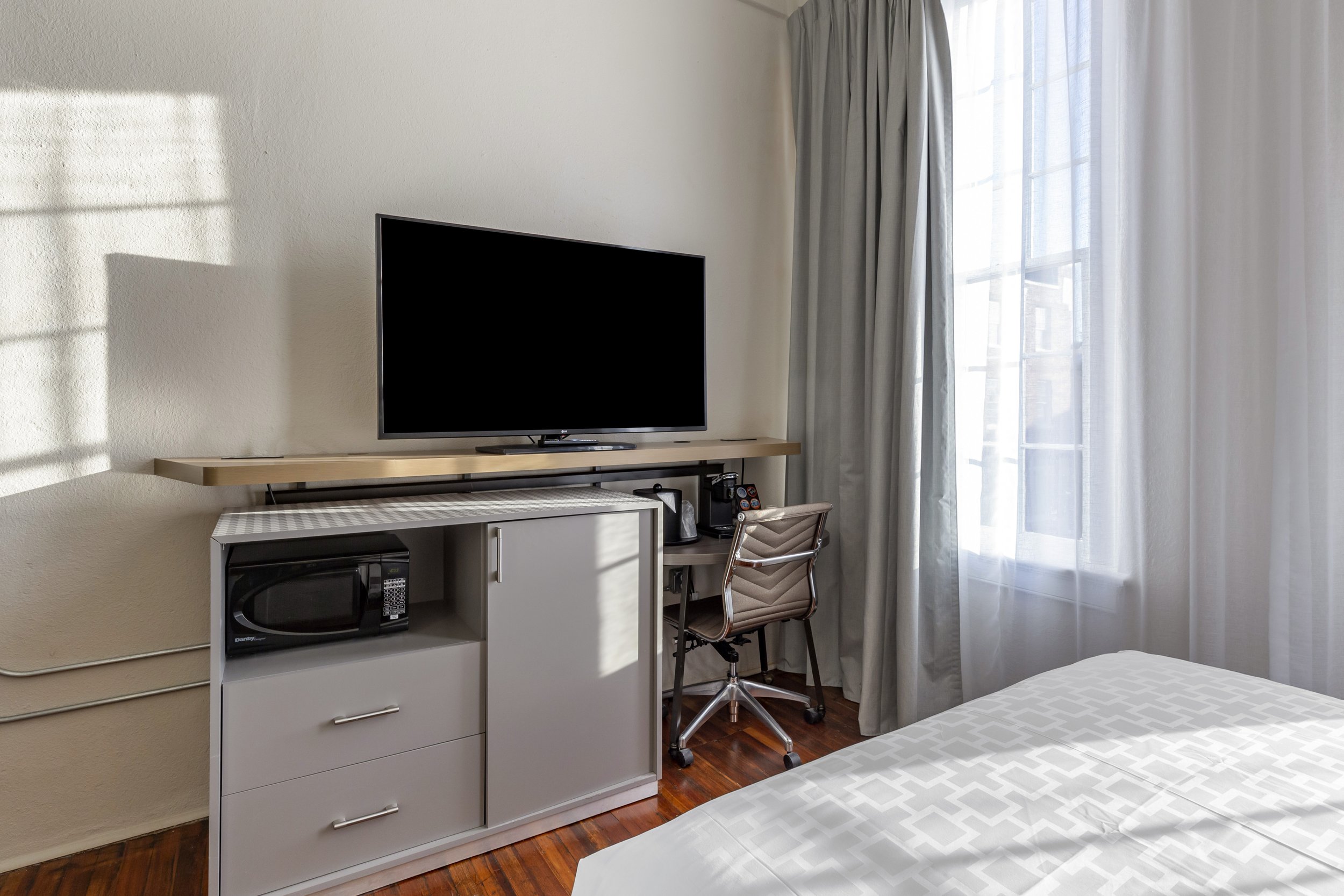 Hotel room with a flat-screen TV, microwave, desk, chair, light-colored curtains, and a window.