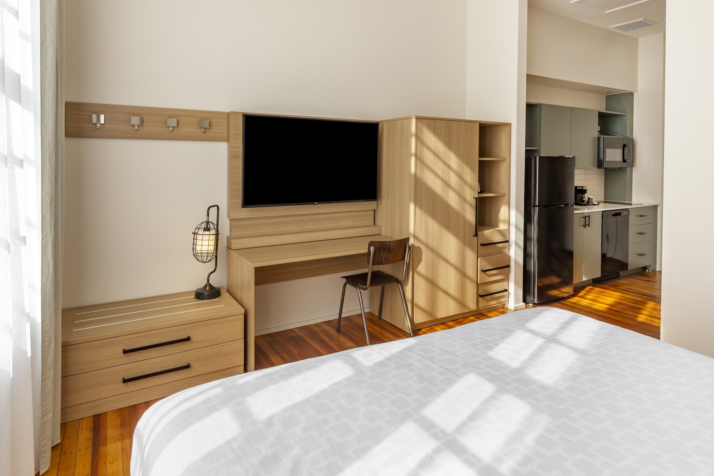 Hotel room with a bed, wooden nightstand, desk, chair, flat-screen TV, kitchenette with refrigerator, microwave, and coffee maker, hardwood floors, sunlight streaming through a window.