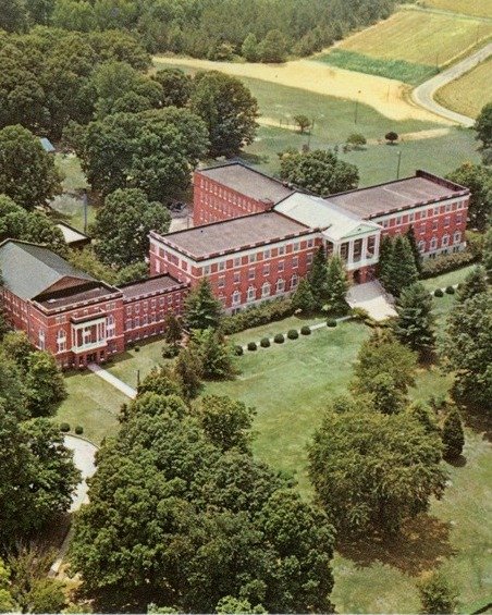 A large, red brick school building with white columns, surrounded by green trees and a manicured lawn.