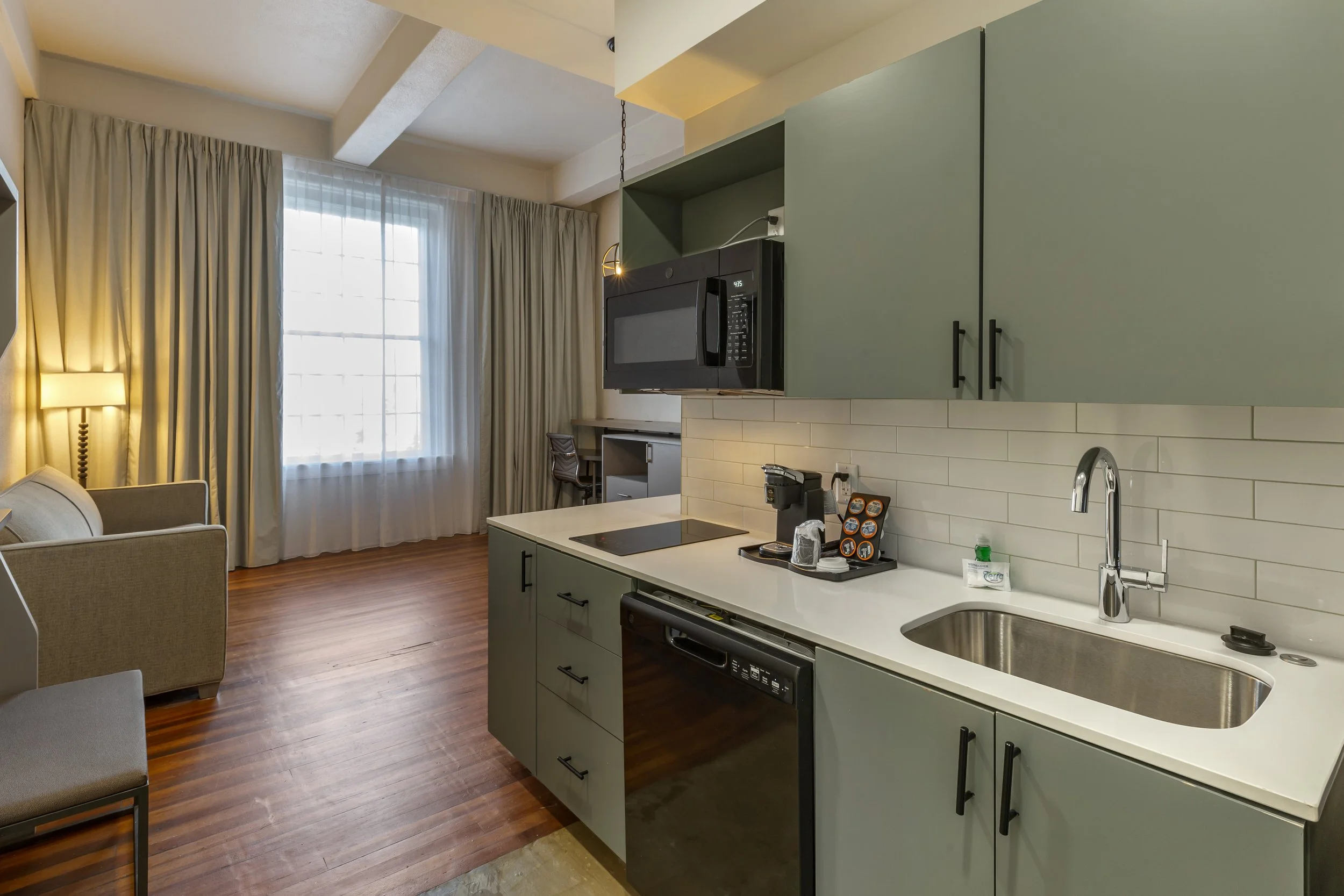 Open-plan studio apartment with a small kitchen area featuring green cabinets, a sink, microwave, coffee maker, and a dishwasher. There is a living space with a beige armchair, a desk, and a large window with white curtains, and hardwood floors throu