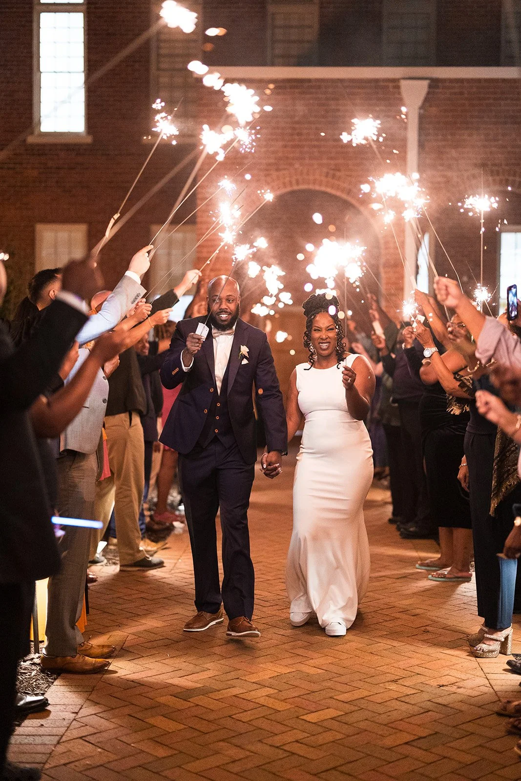 A bride and groom walking hand in hand through a crowd holding sparklers at night, celebrating their wedding with bright smiles.