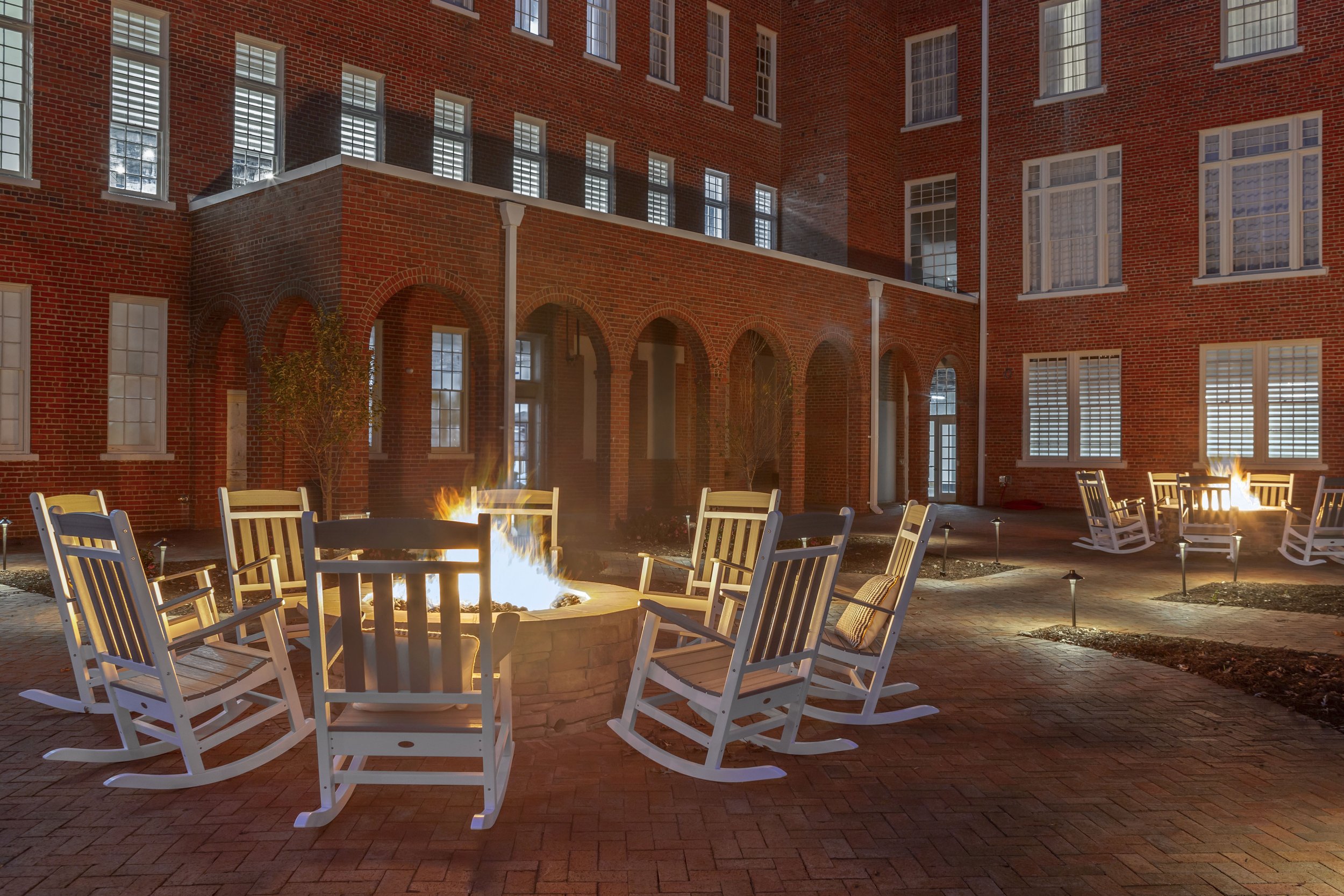 Outdoor courtyard at night with brick buildings, lit by fire pits surrounded by white rocking chairs and small landscape lights.