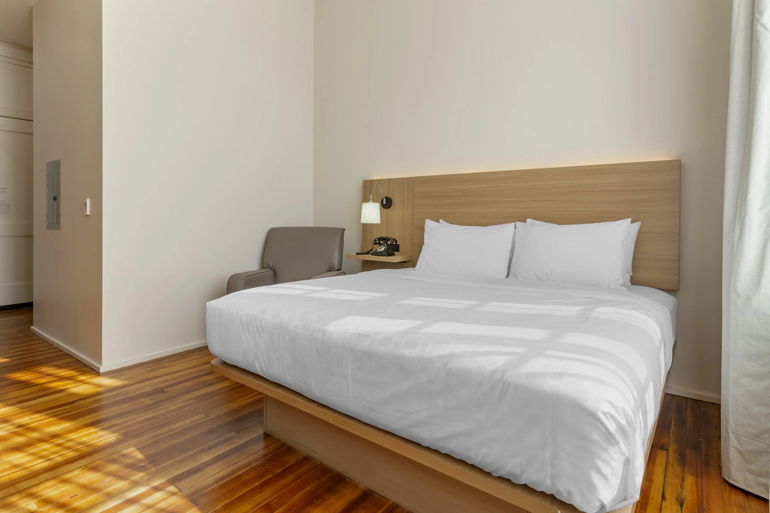 Hotel room with white bed, wooden headboard, grey armchair, wall-mounted lamp, vintage black rotary phone, and light-colored curtains.