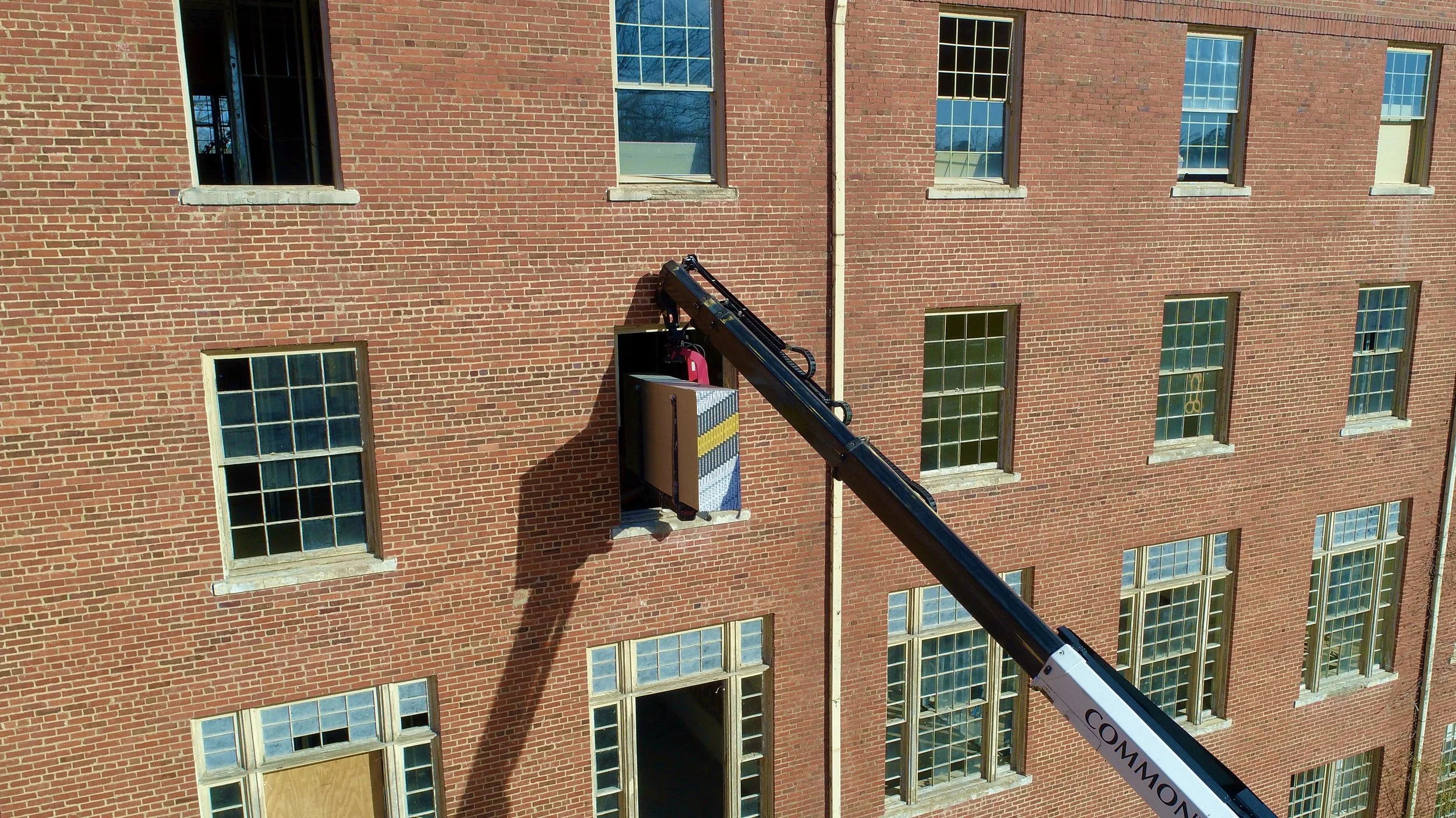 A crane with a long black arm is lifting an HVAC unit on the side of a red brick building with multiple windows.