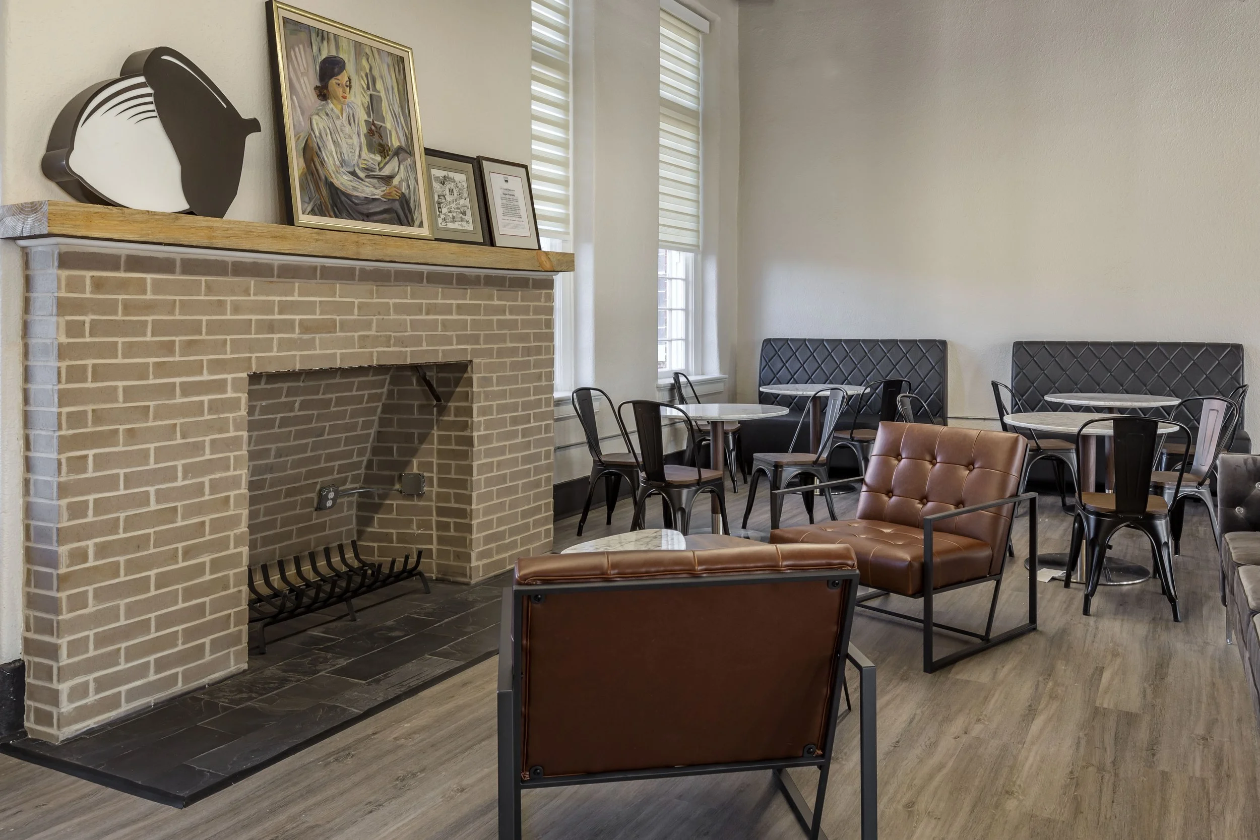 Interior of a cozy cafe with a brick fireplace, modern chairs, and tables, decorated with art and framed pictures on the mantel.