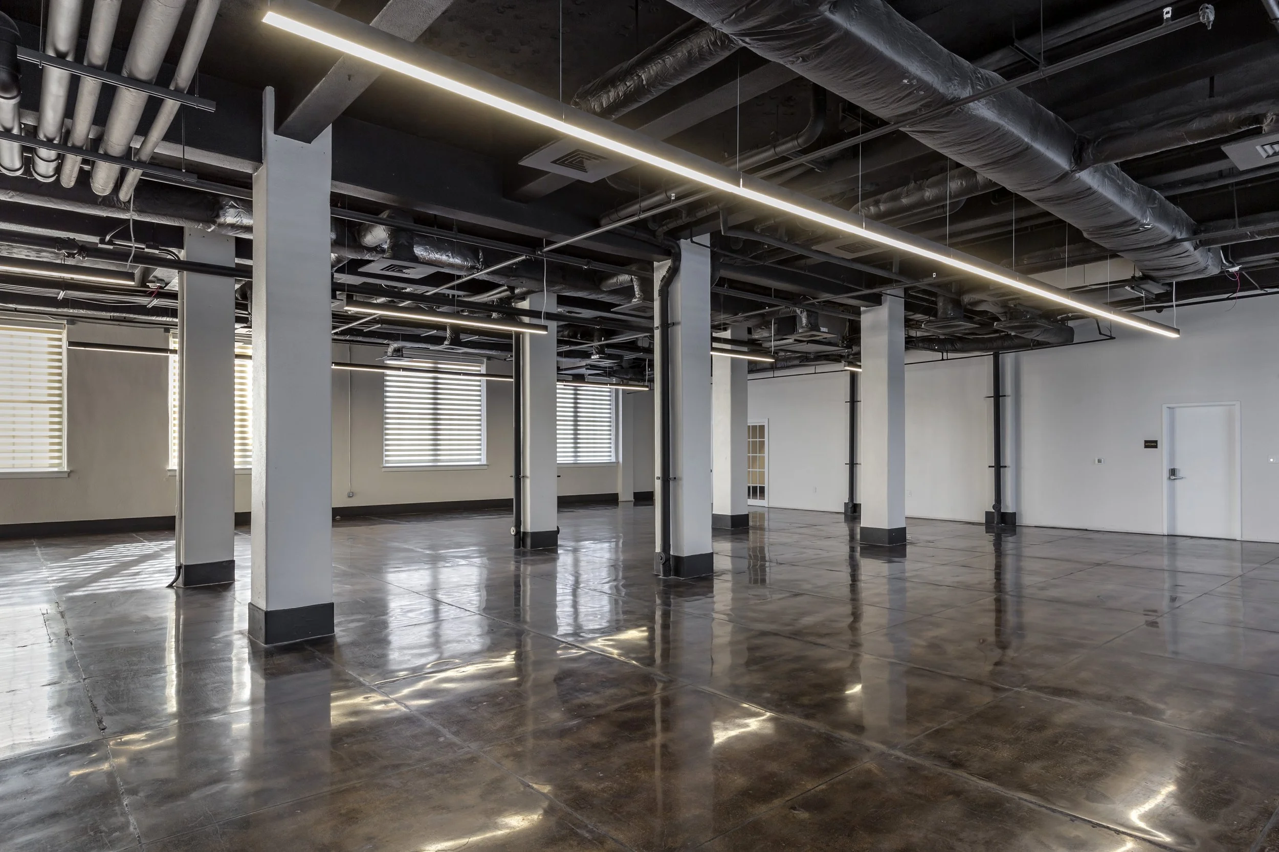 Empty commercial space with polished concrete floors, white walls, black ceiling with exposed pipes and ducts, large windows with blinds, and modern linear lighting.