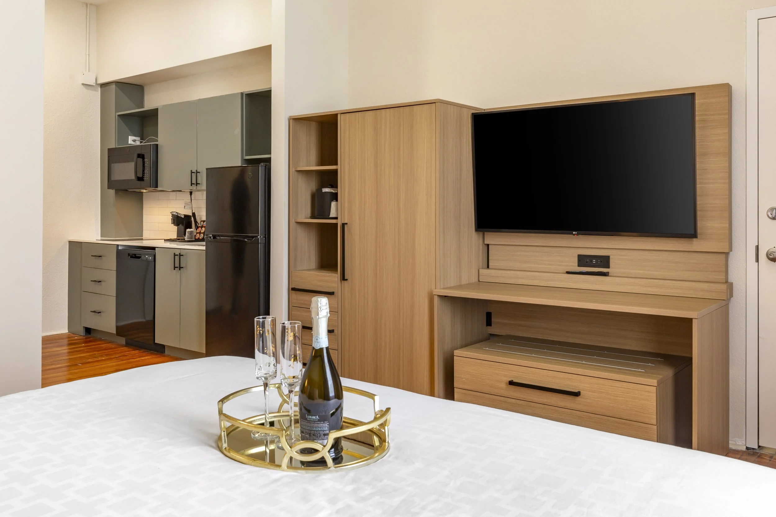 Kitchen area with black refrigerator, microwave, coffee maker, and kitchen cabinets in the background, with a wooden TV stand and flat-screen TV in the foreground, and a bottle of champagne with two glasses on a tray on the bed.
