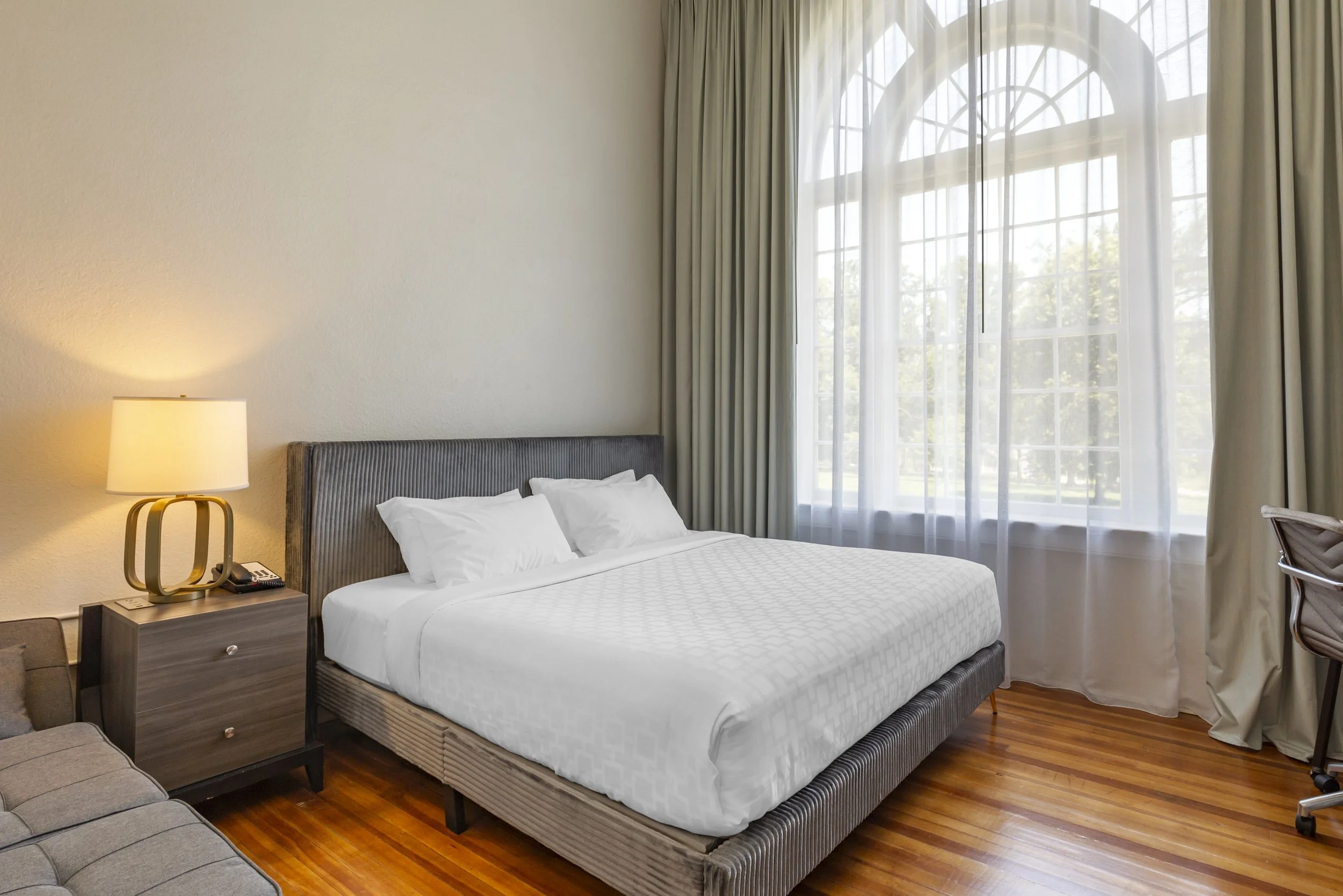 A bedroom with a large bed with white pillows and a textured white duvet, a nightstand with a modern lamp, a gray upholstered headboard, hardwood floors, and a large window with sheer and heavy curtains.