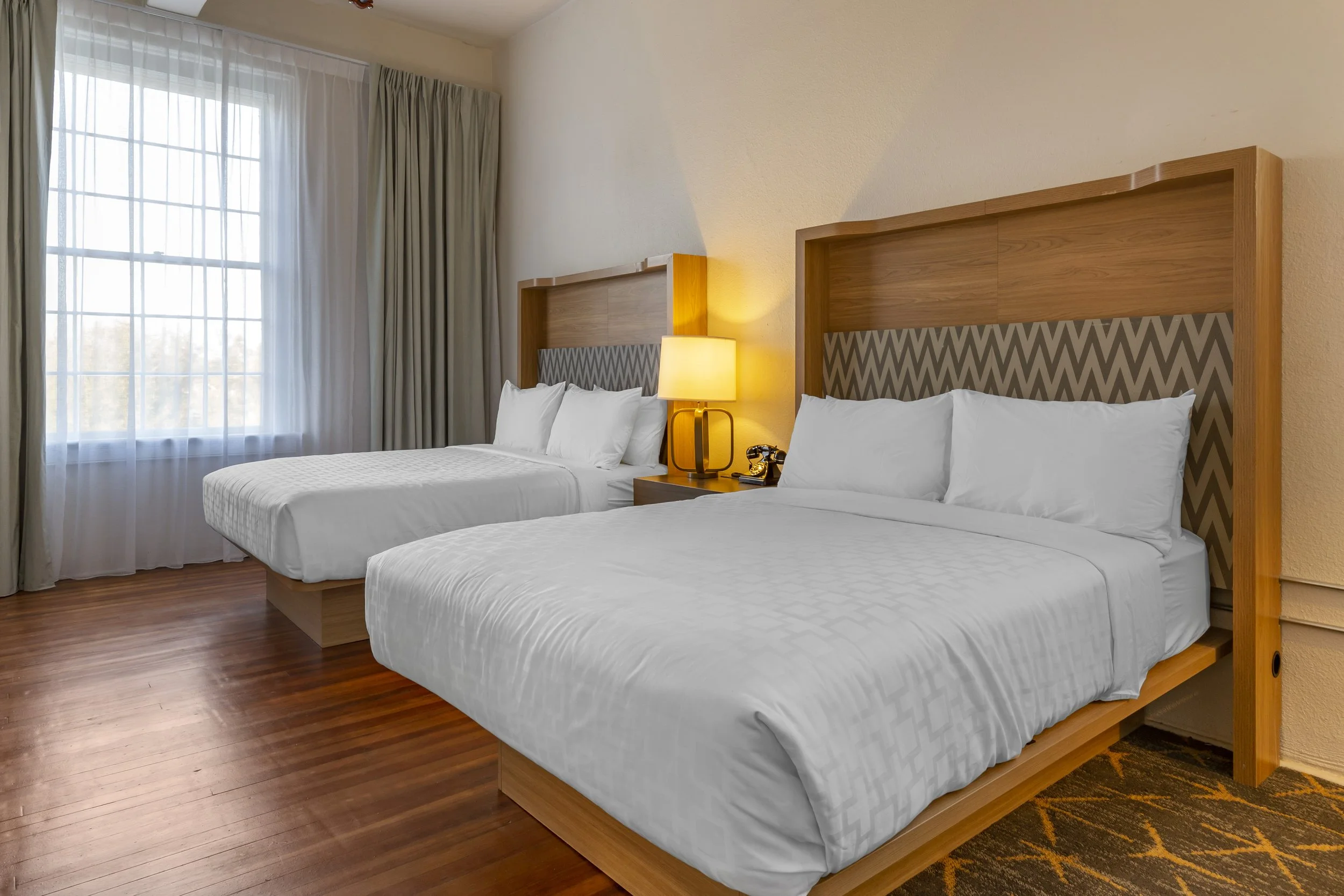 Two double beds with white linens and headboards, a nightstand with a lamp and a vintage phone, window with curtains, wooden flooring, and partial view of patterned carpet.