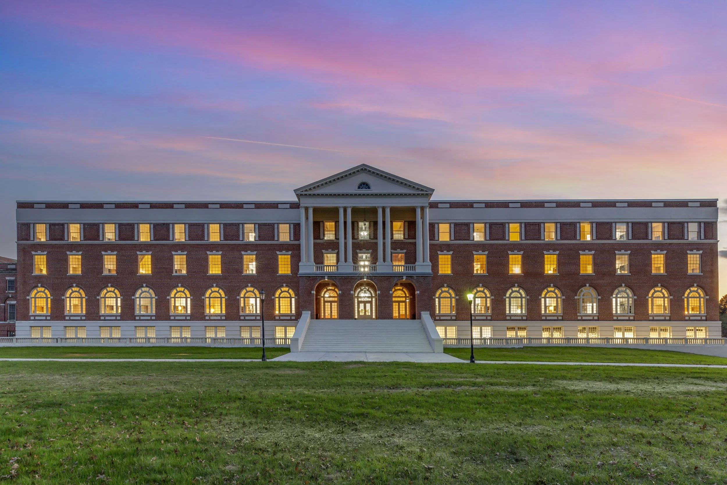 A large brick building with classical architecture, tall columns at the entrance, multiple lit windows, and a staircase leading up to the main door, under a colorful sunset sky.
