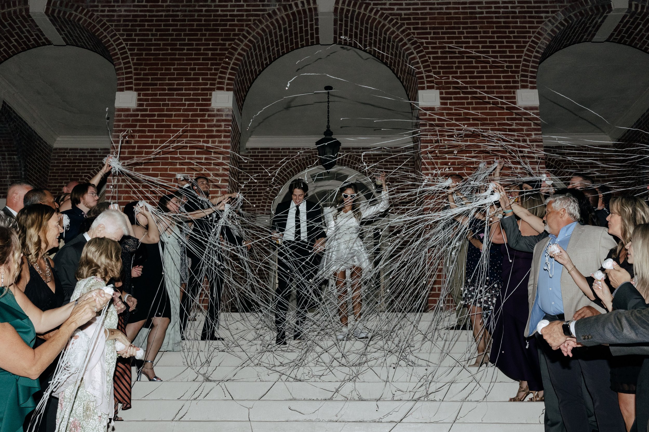 A bride and groom exiting a building as guests throw streamers on the stairs.