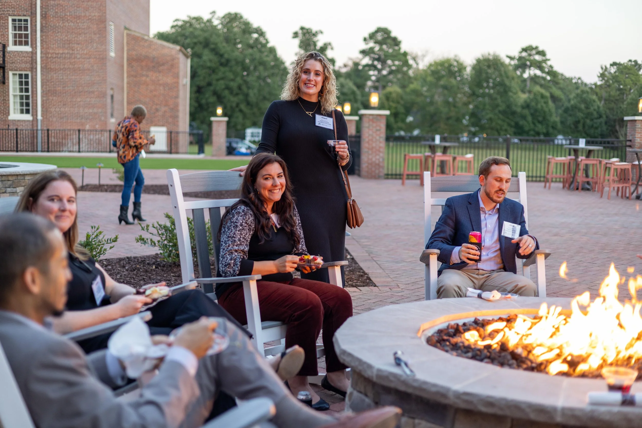 A group of people enjoying an outdoor gathering around a fire pit, with some holding drinks and snacks, on a patio with chairs and brick building in the background.