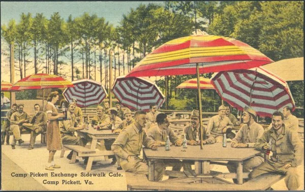 A vintage scene at Camp Pickett in Virginia shows soldiers sitting and standing around picnic tables under large, colorful striped umbrellas. The background features trees and a blue sky, with additional umbrellas and soldiers in the distance.