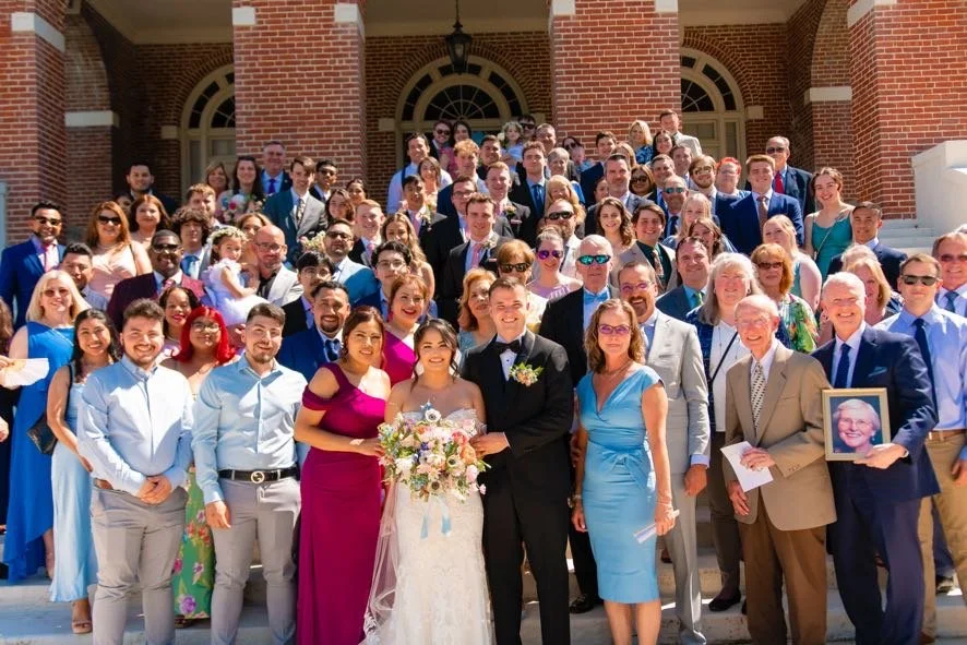 Large wedding photo of a bride and groom with family and friends on steps outside a brick building.