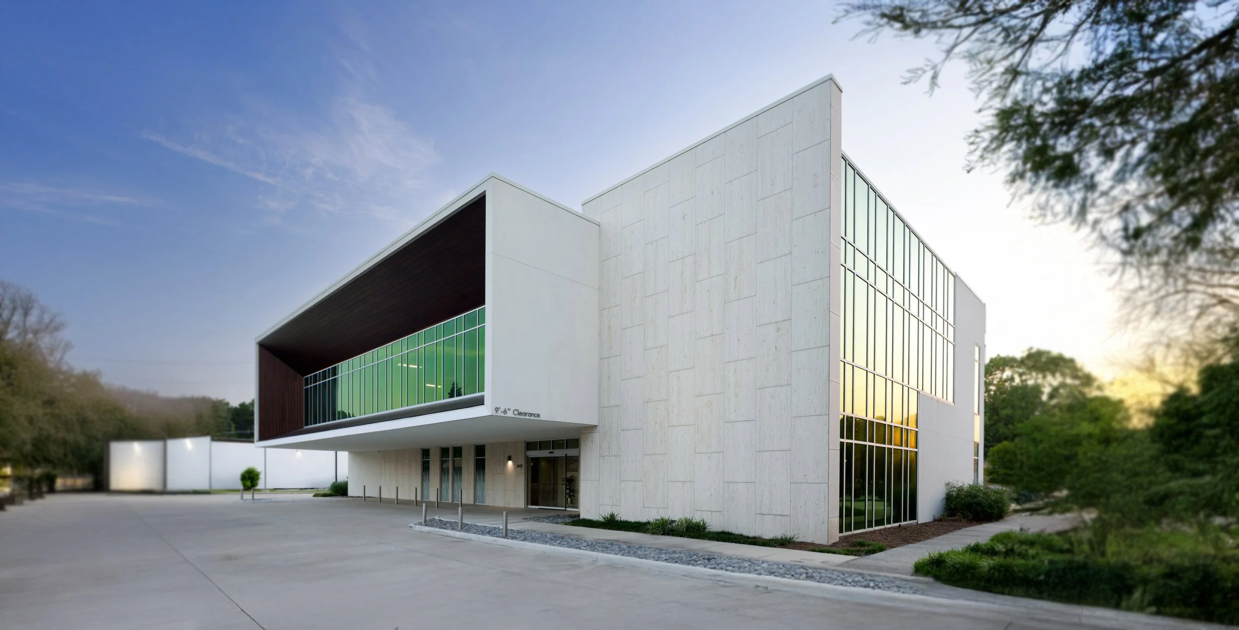 Modern white building with large glass windows and a unique architectural design, surrounded by greenery and a clear sky at sunset.