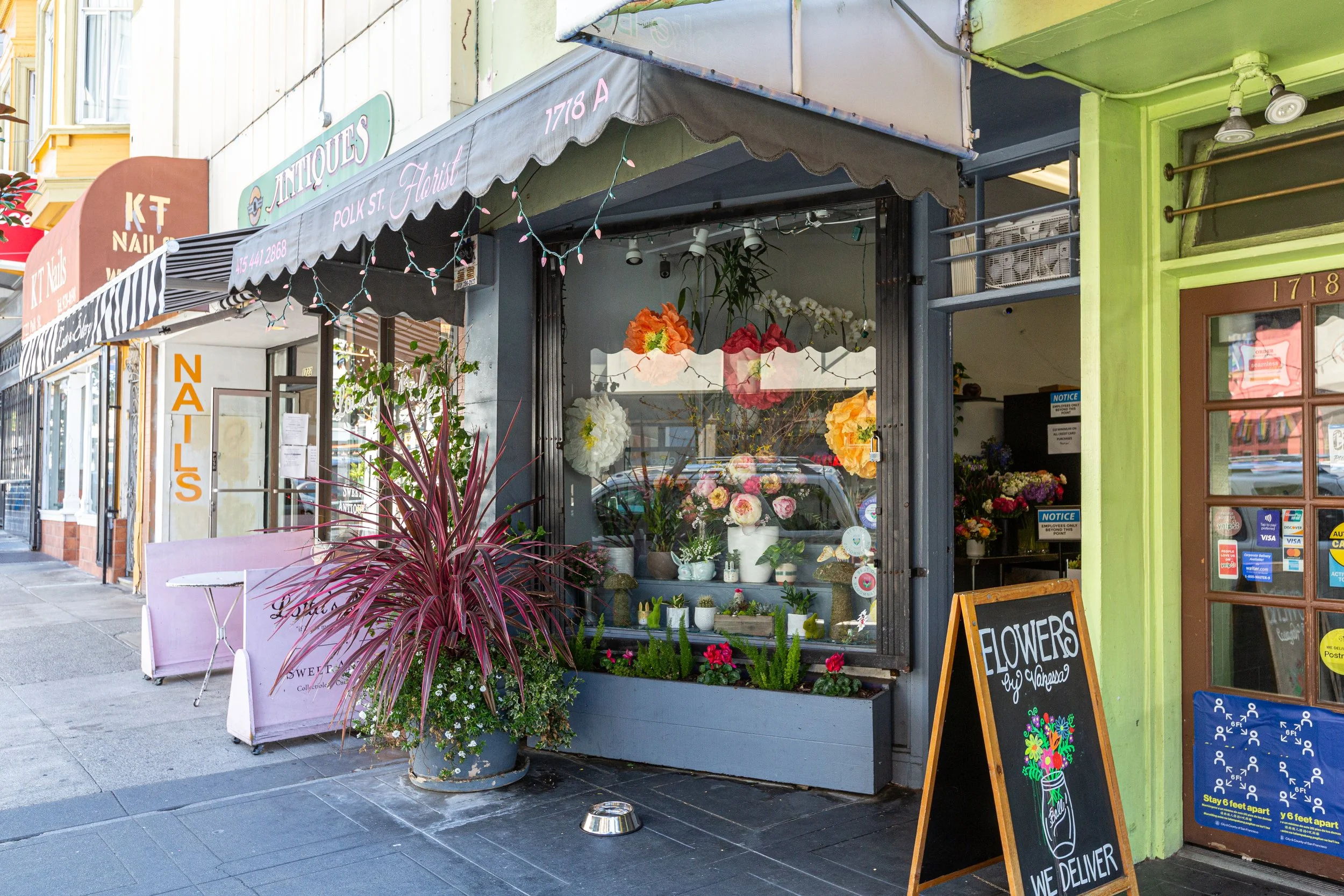 A flower shop with a glass window display of colorful flowers and plants. Outside, there is a large potted pink and green plant, a chalkboard sign with a flower illustration and the words 'Flowers by Noreva' and 'We Deliver', and a pink bench. The sh