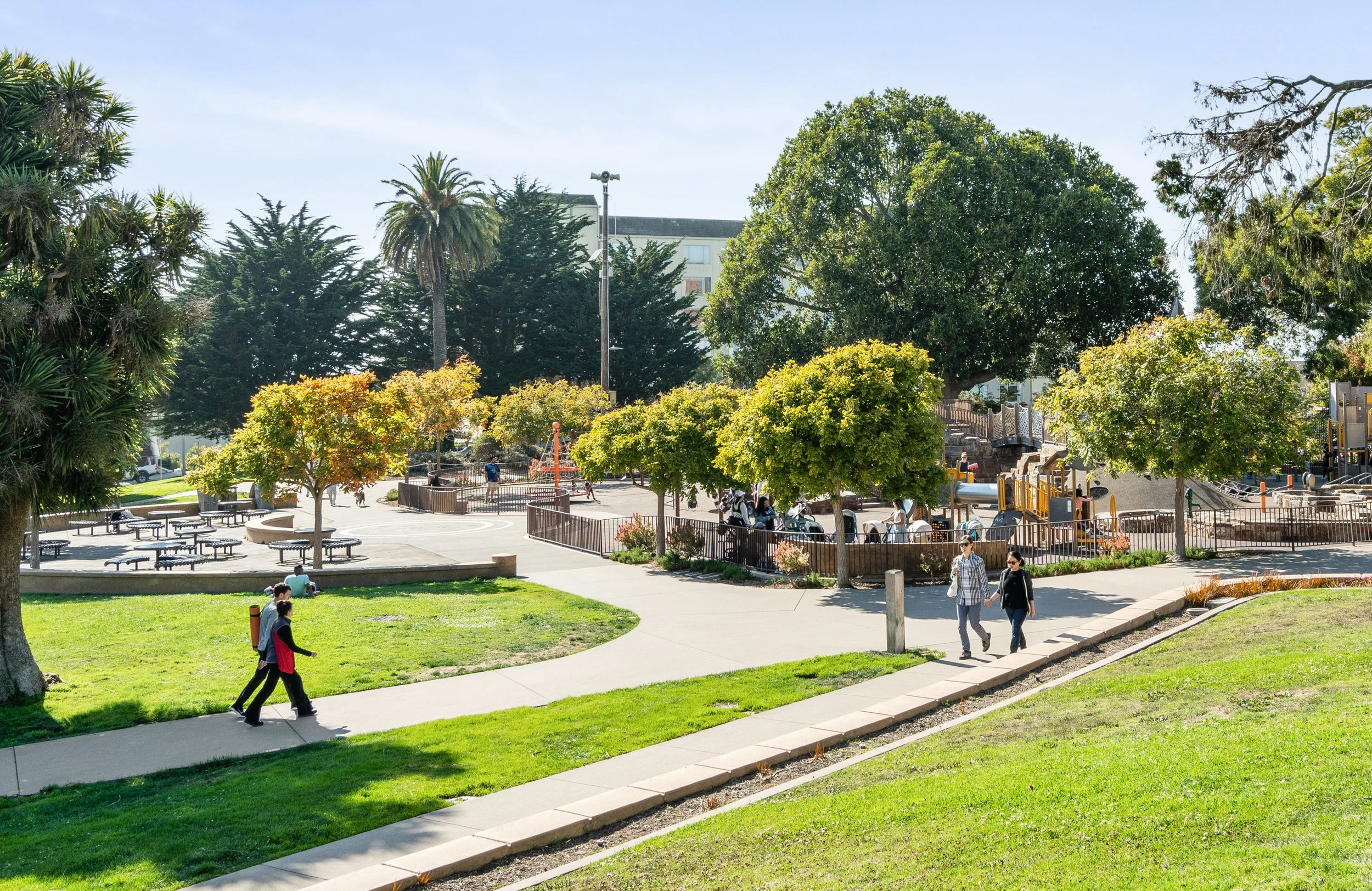 People walking and sitting in a park with trees, grassy areas, and a playground.